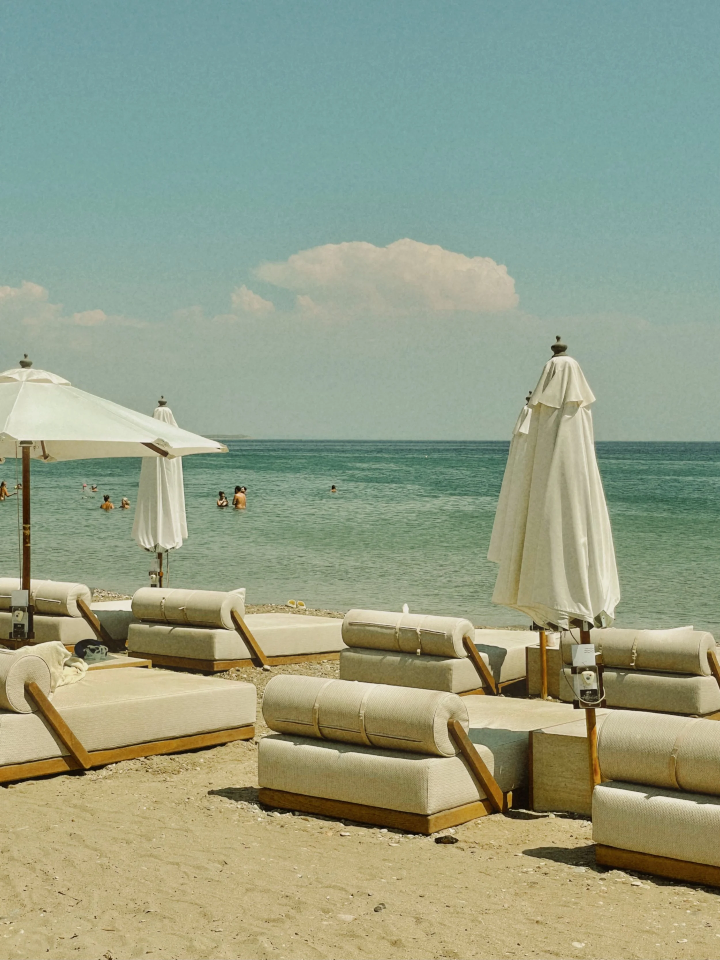 Empty beach lounge chairs with umbrellas facing the ocean on a sandy beach with people swimming in the water and a partly cloudy sky.