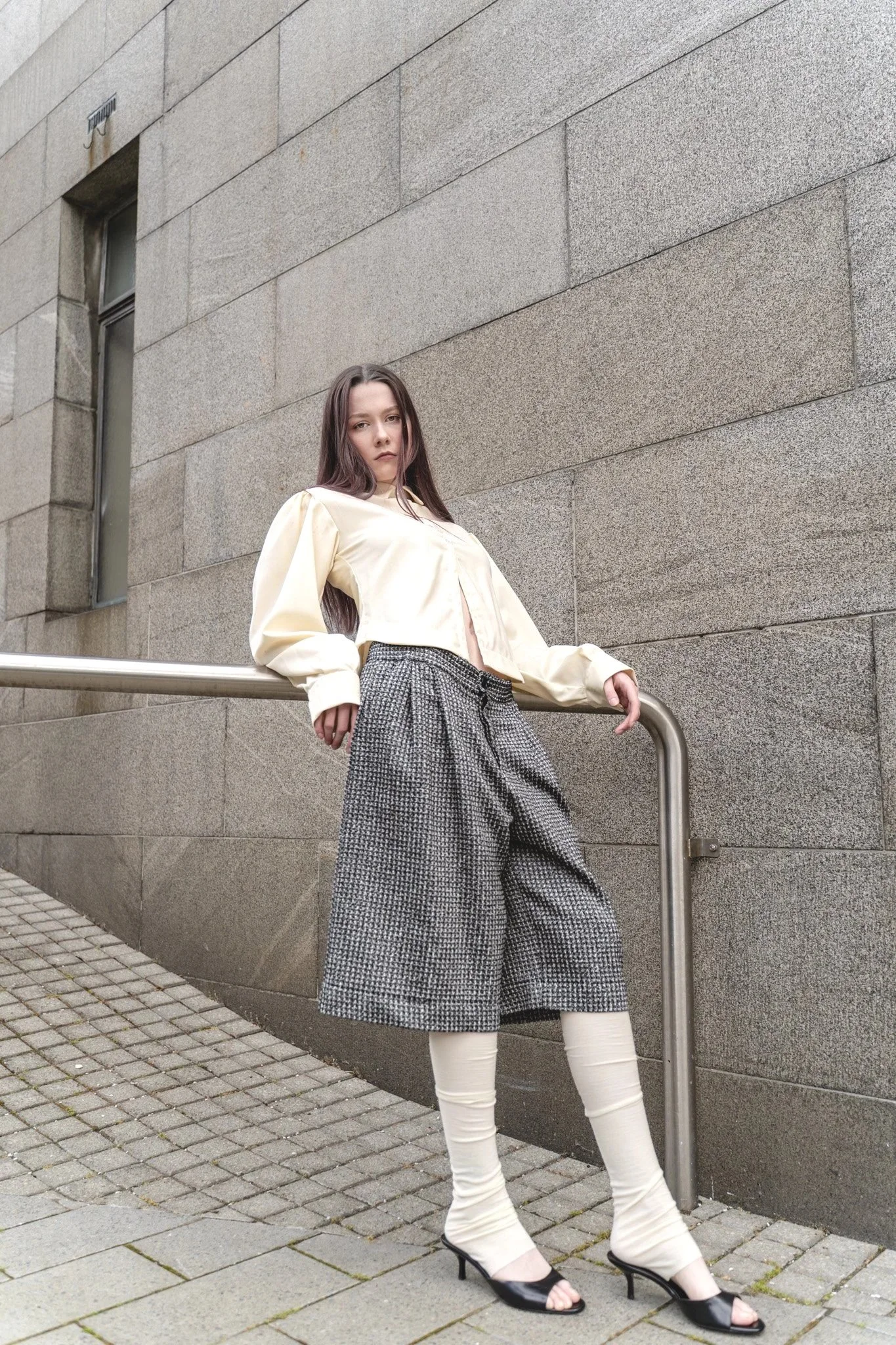A woman standing outdoors against a stone wall, wearing a cream jacket, plaid wide-leg pants, cream leg warmers, and black open-toe heels.