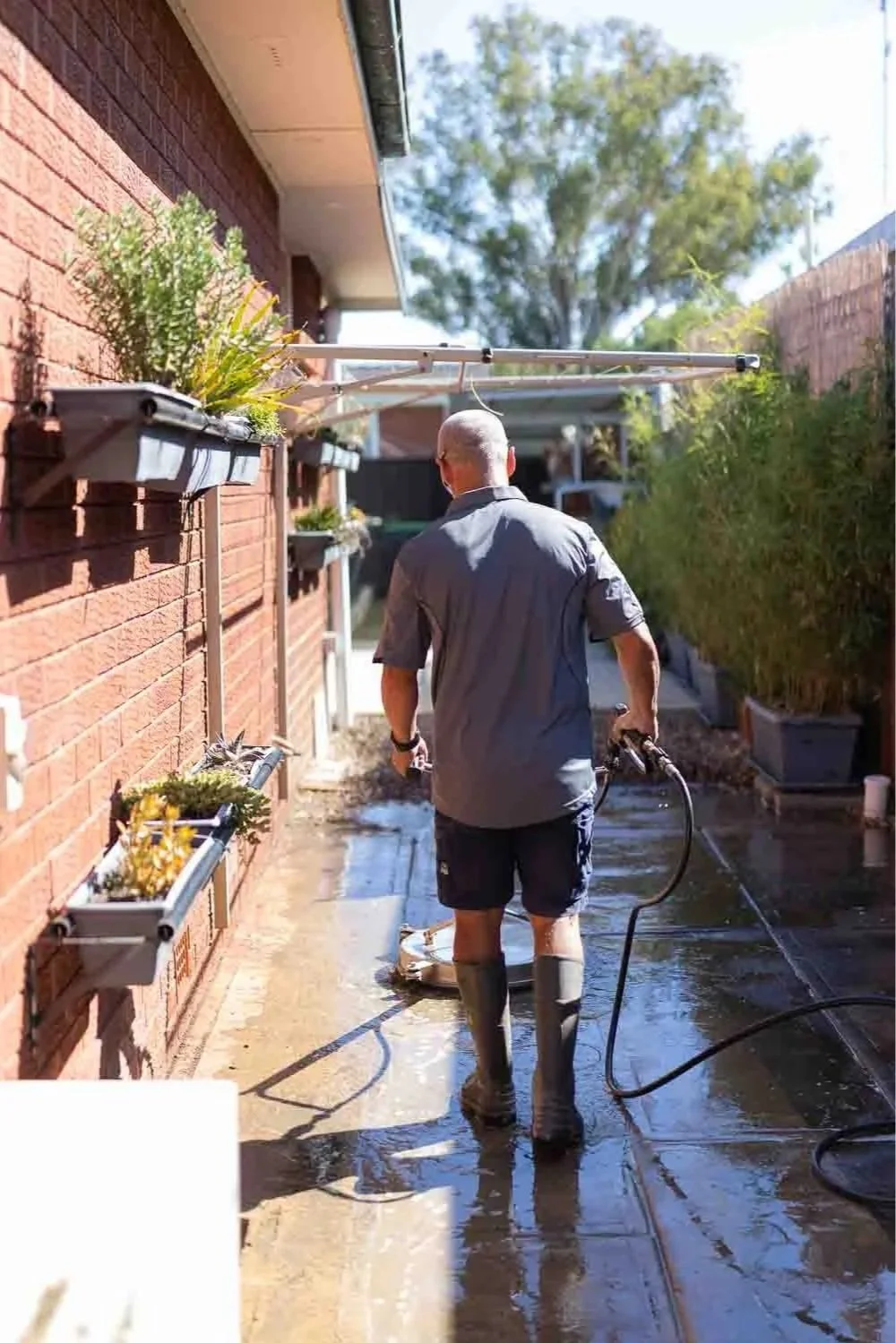 A man wearing rubber boots and casual clothes is pressure washing a patio outside a brick house on a sunny day.