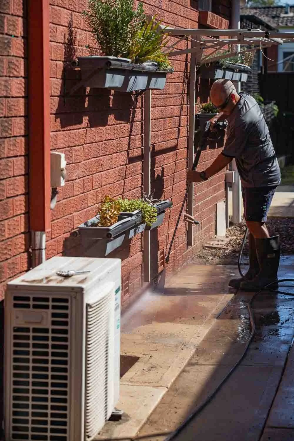 A person using a pressure washer to clean the sidewalk beside a brick wall with hanging planters and an outdoor air conditioning unit.