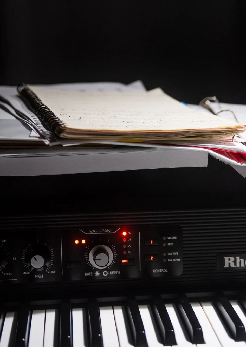 This is a Rhodes piano built in 2024, the Mark 8, that is photographed with a black background and the vari-pan visible above the black and white keys (the LED lights are beautifully turned on and are in shades of orange), with a binder of music abov
