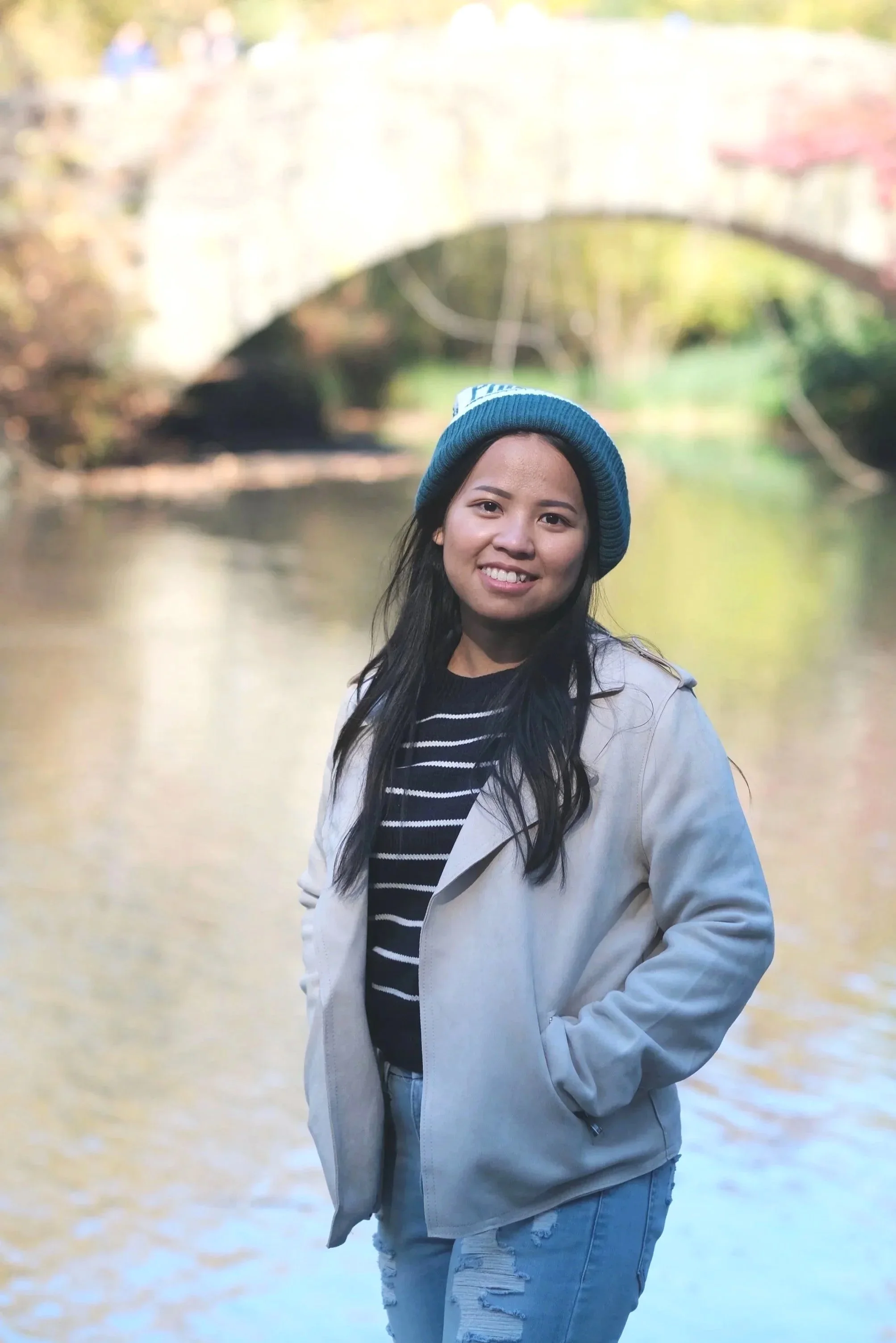 A young woman with long dark hair, wearing a blue beanie, black and white striped shirt, light-colored jacket, and ripped jeans, standing by a river with a stone bridge in the background.