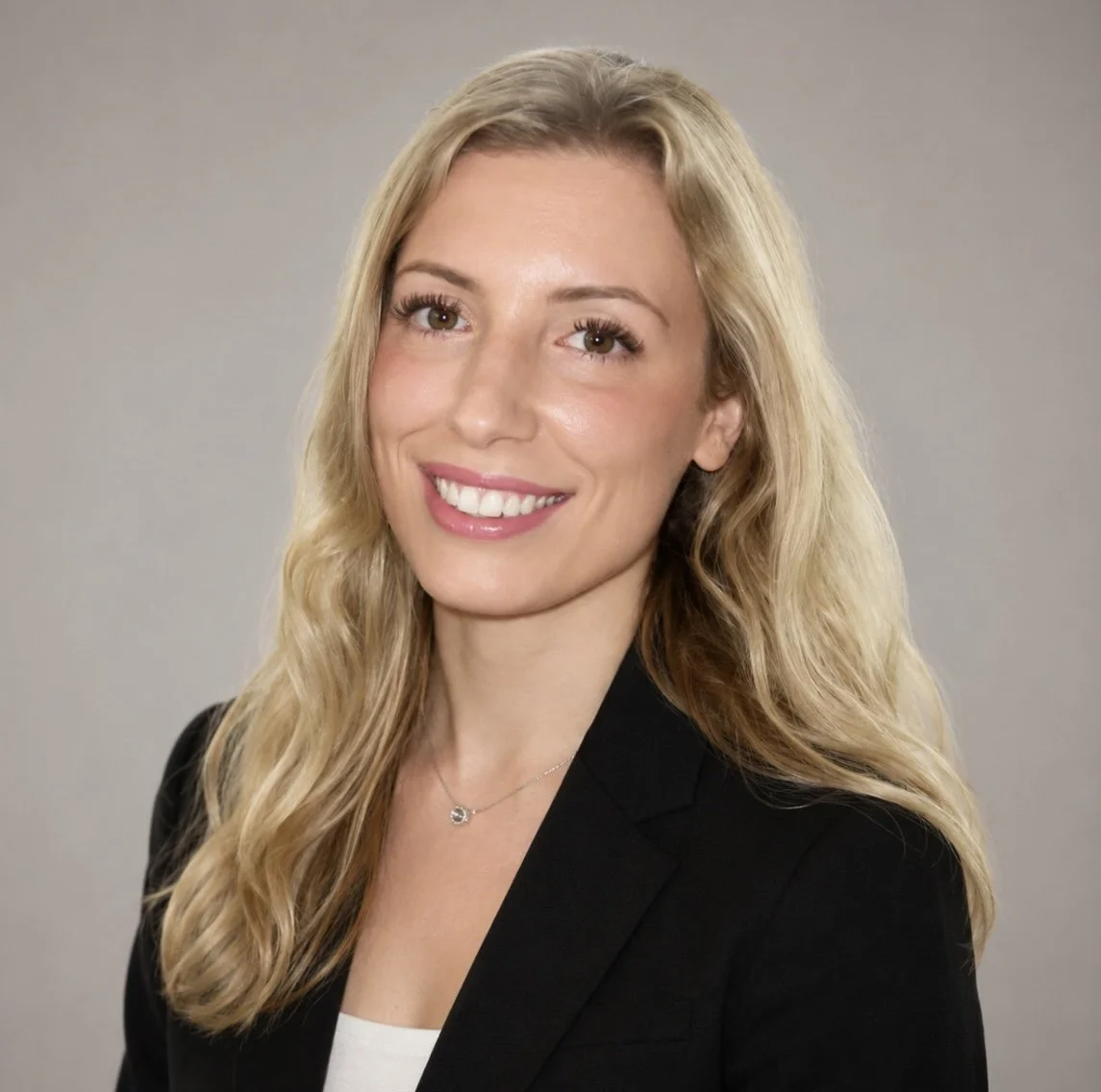 A professional woman with long blonde hair, smiling, wearing a black blazer and a necklace, against a neutral gray background.