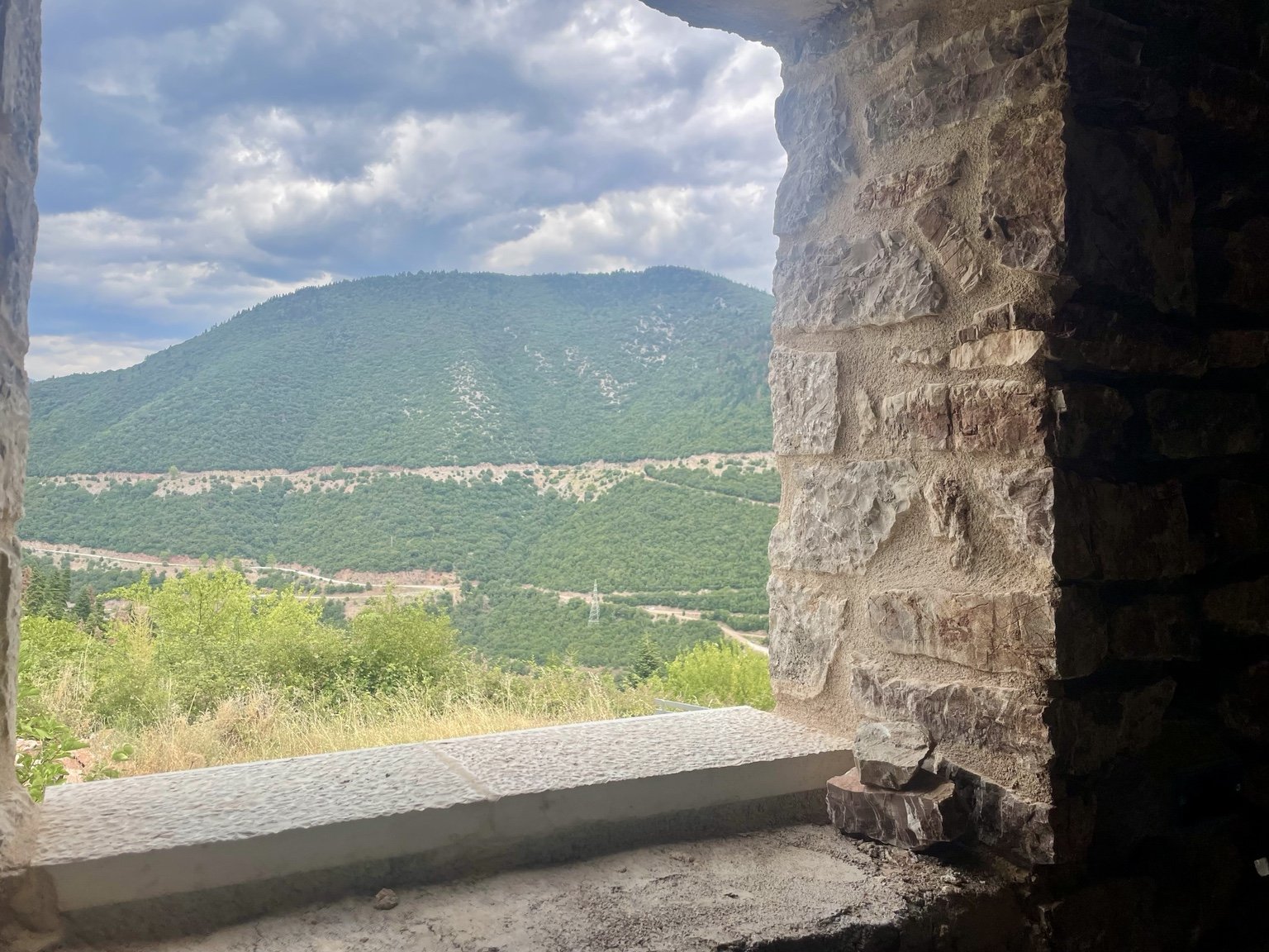 View through a stone window opening showing a lush green mountain landscape with a cloudy sky.