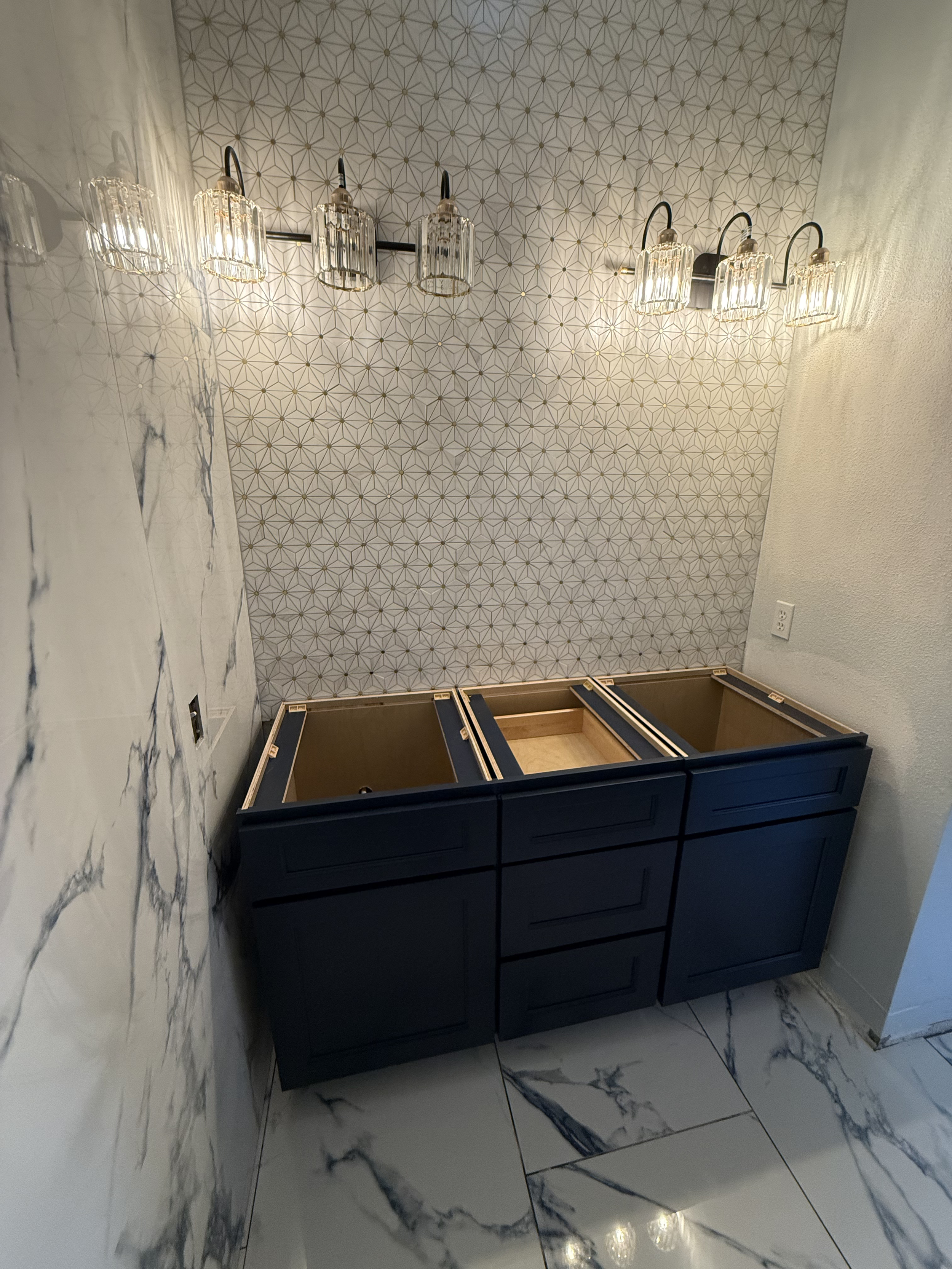 Unfinished navy blue cabinet with drawers on a marble floor, mounted on a wall with patterned wallpaper and a marble wall on the side, illuminated by a modern chandelier with exposed bulbs.
