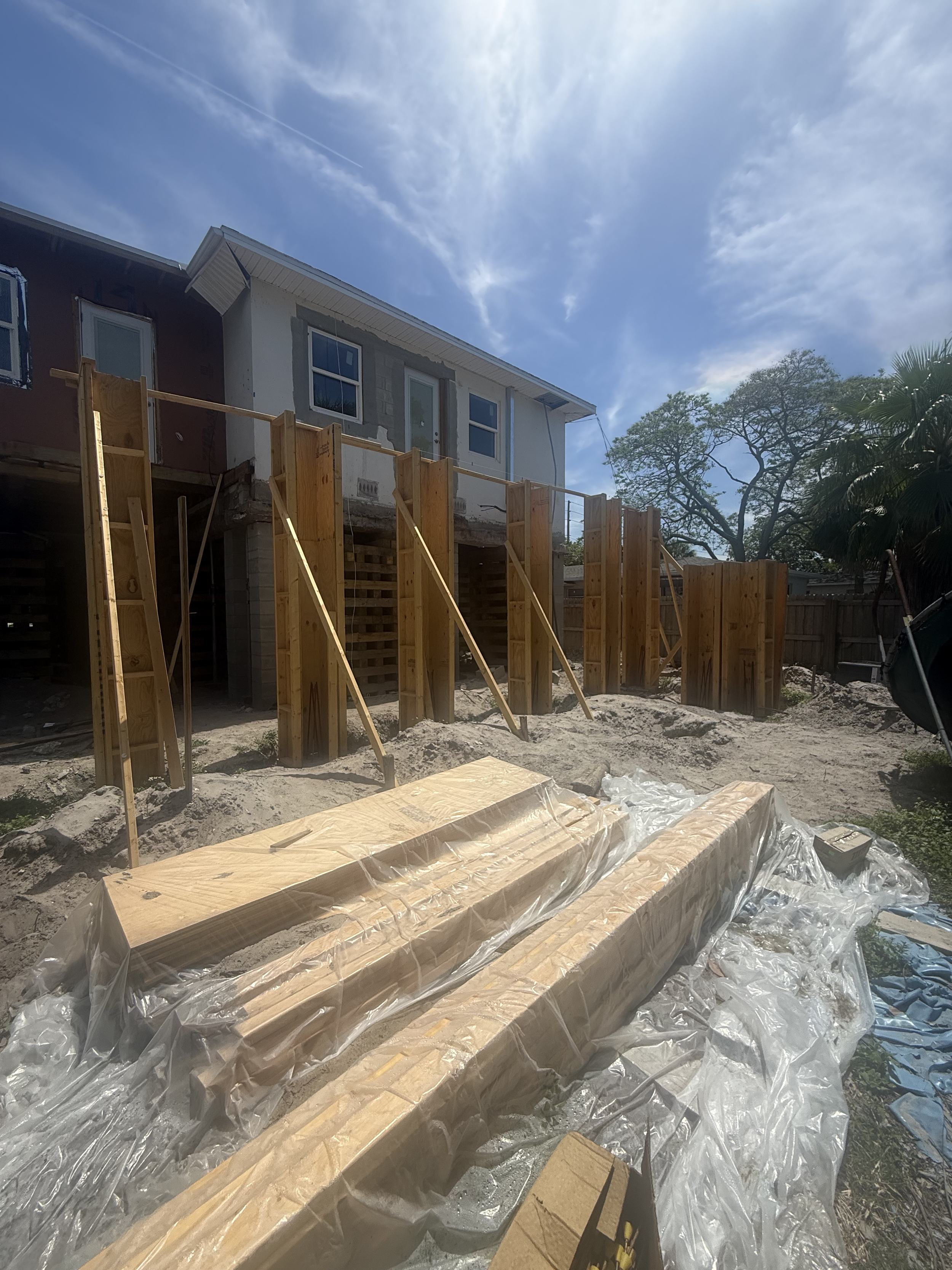 A house under construction with exposed framing, construction materials, and plastic sheeting on the ground in the foreground. The sky is partly cloudy with sunlight shining through.