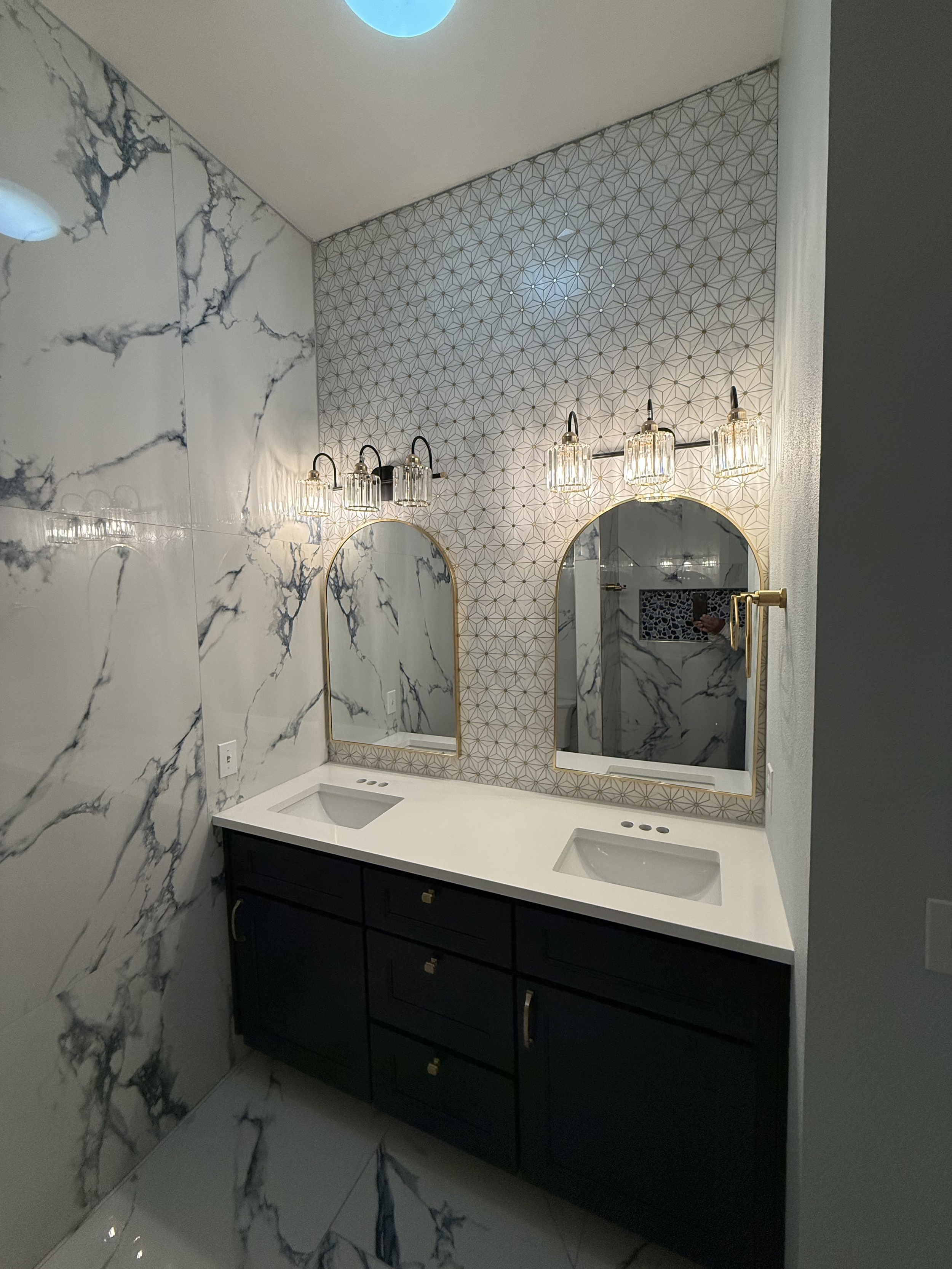 A modern bathroom with white marble walls and floors, two mirrors with gold trim above a black vanity with two white sinks, and decorative wall lights above the mirrors.