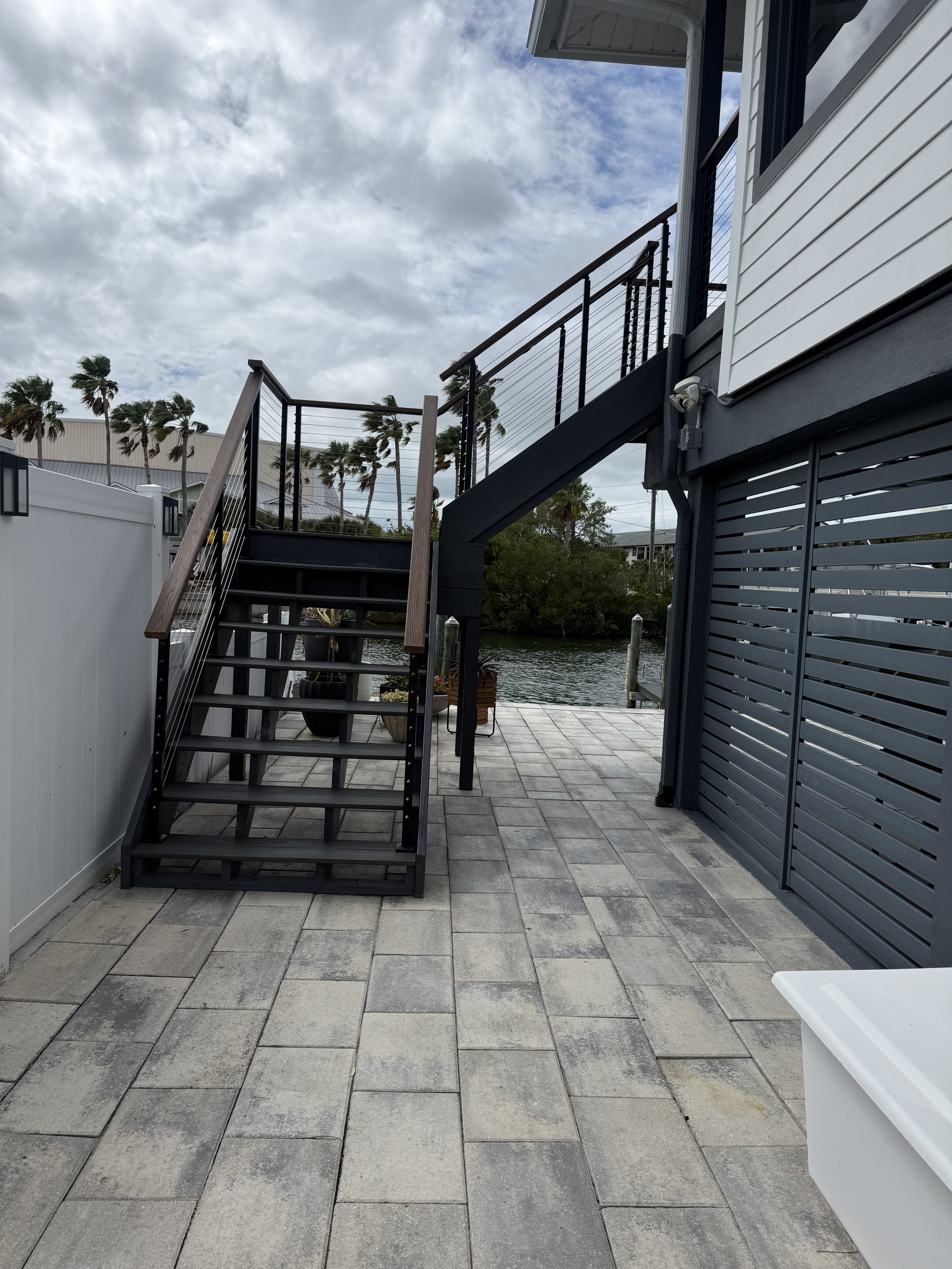 Outdoor patio area with tiled flooring, metal stairs leading to an upper deck with a black railing, a white fence on the left, and a house with white siding on the right. There is a view of palm trees and water in the background, under cloudy sky.