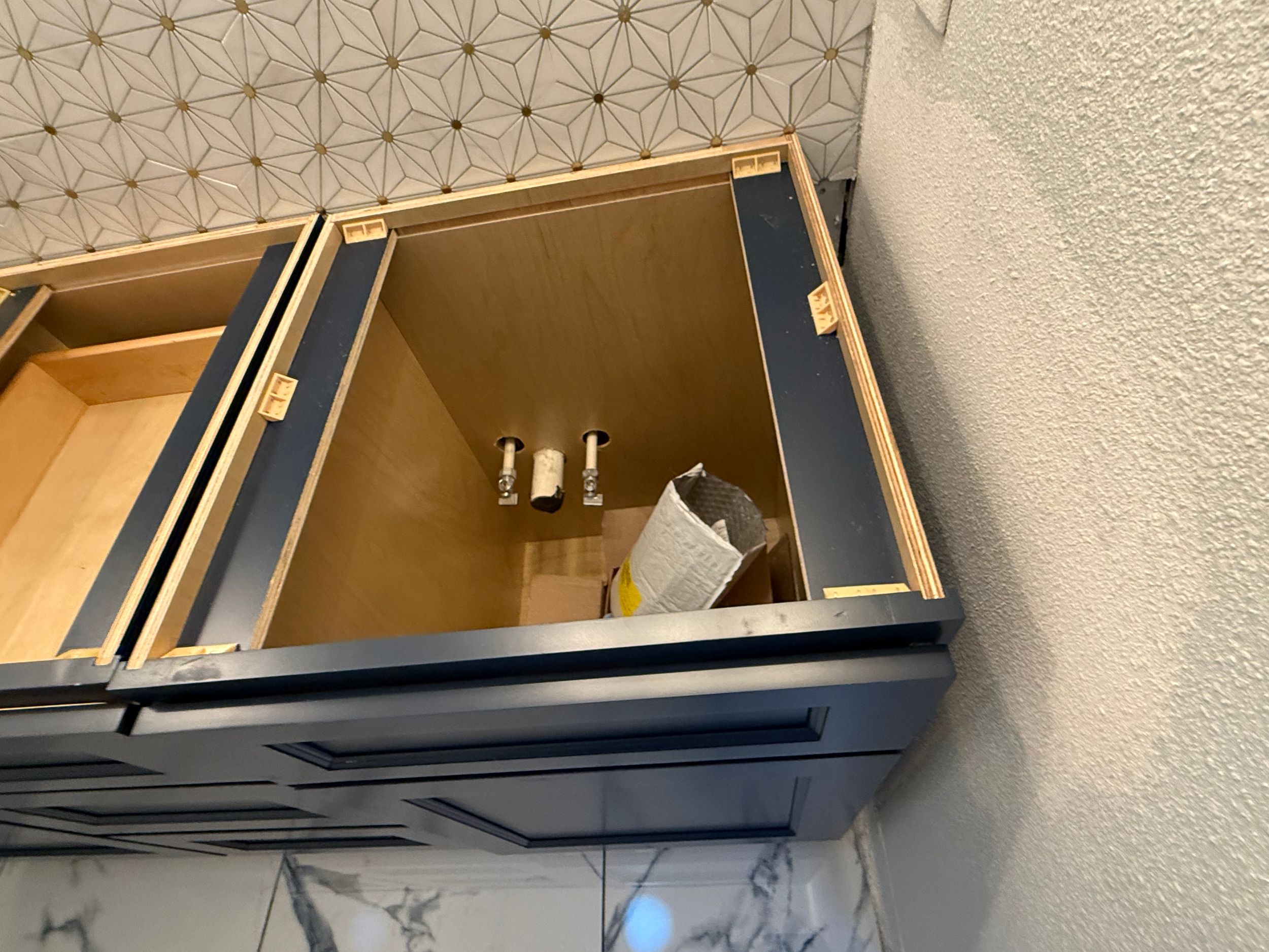 Open cabinet with black and wood framework, interior pipes, and a bag inside. Wall with geometric pattern wallpaper and a textured ceiling.