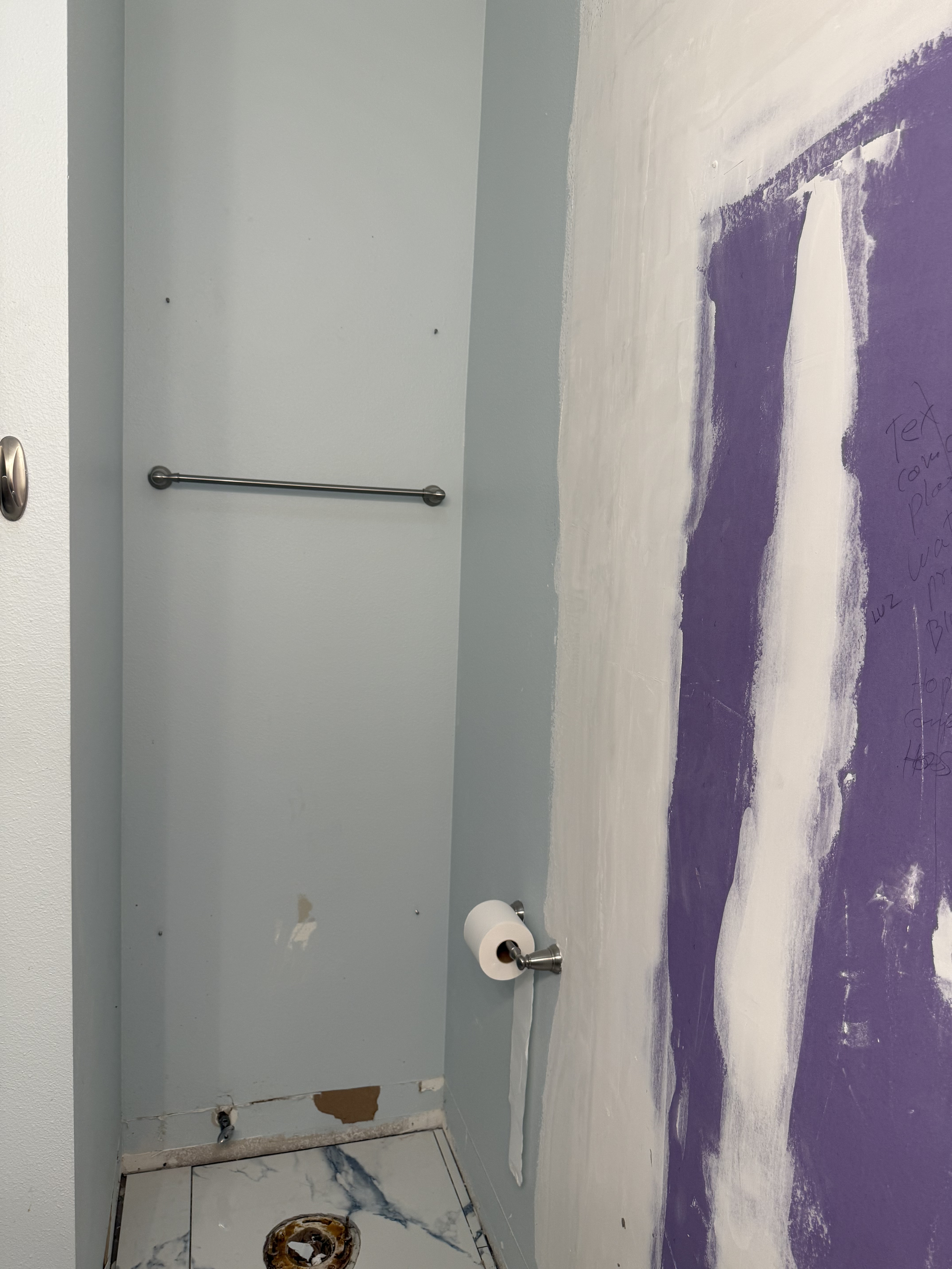 Interior of a room under renovation with a partially painted purple and white wall, a white towel bar, a partially visible toilet paper holder, a bit of damaged wall with peeling paint and exposed drywall, and a marble tile floor.