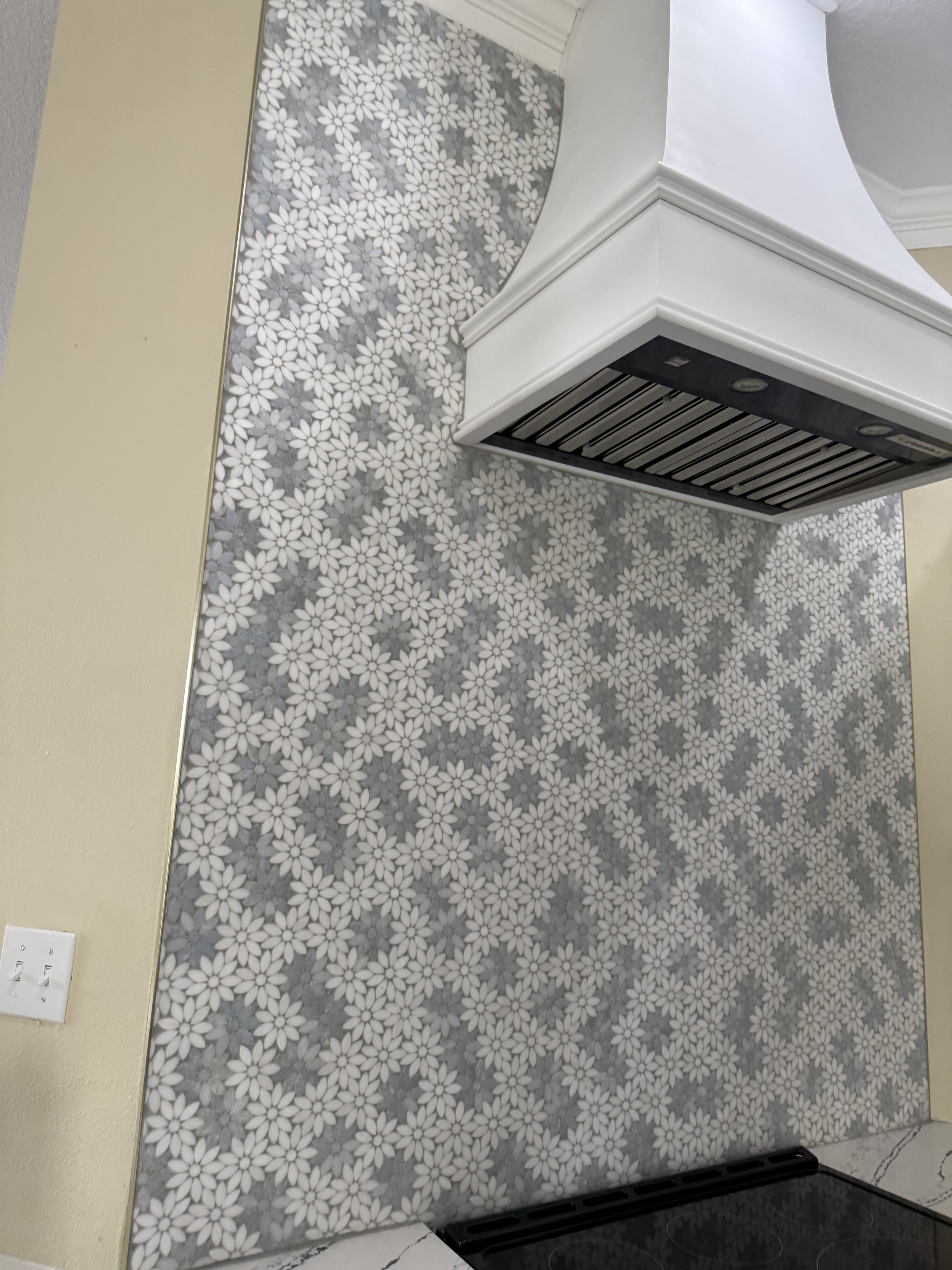 Wall with decorative gray and white mosaic tile featuring white daisy flower patterns, next to a light switch and kitchen vent.