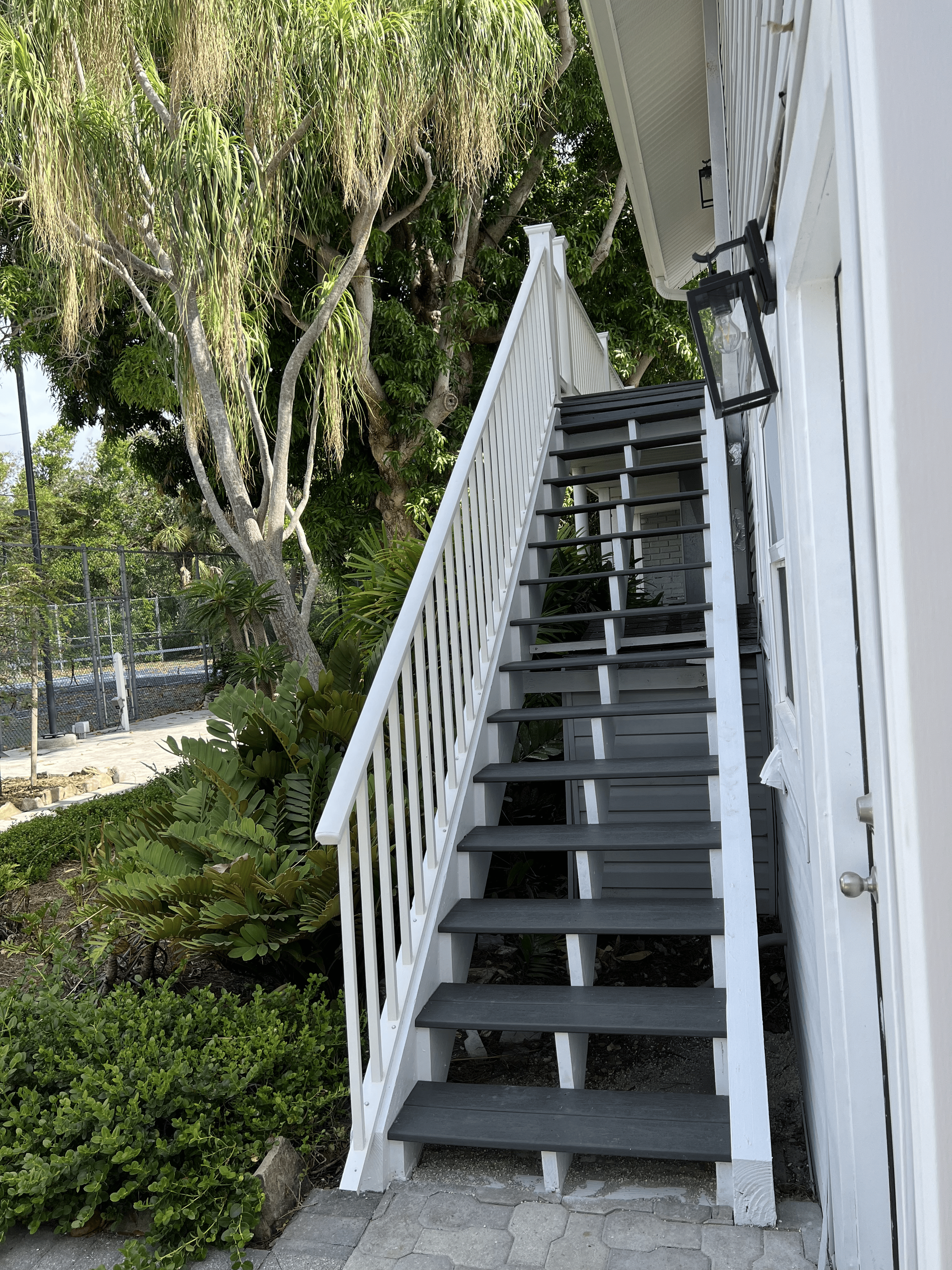 Exterior staircase with white railings and black steps leading to a second-floor entrance, surrounded by green plants and trees.