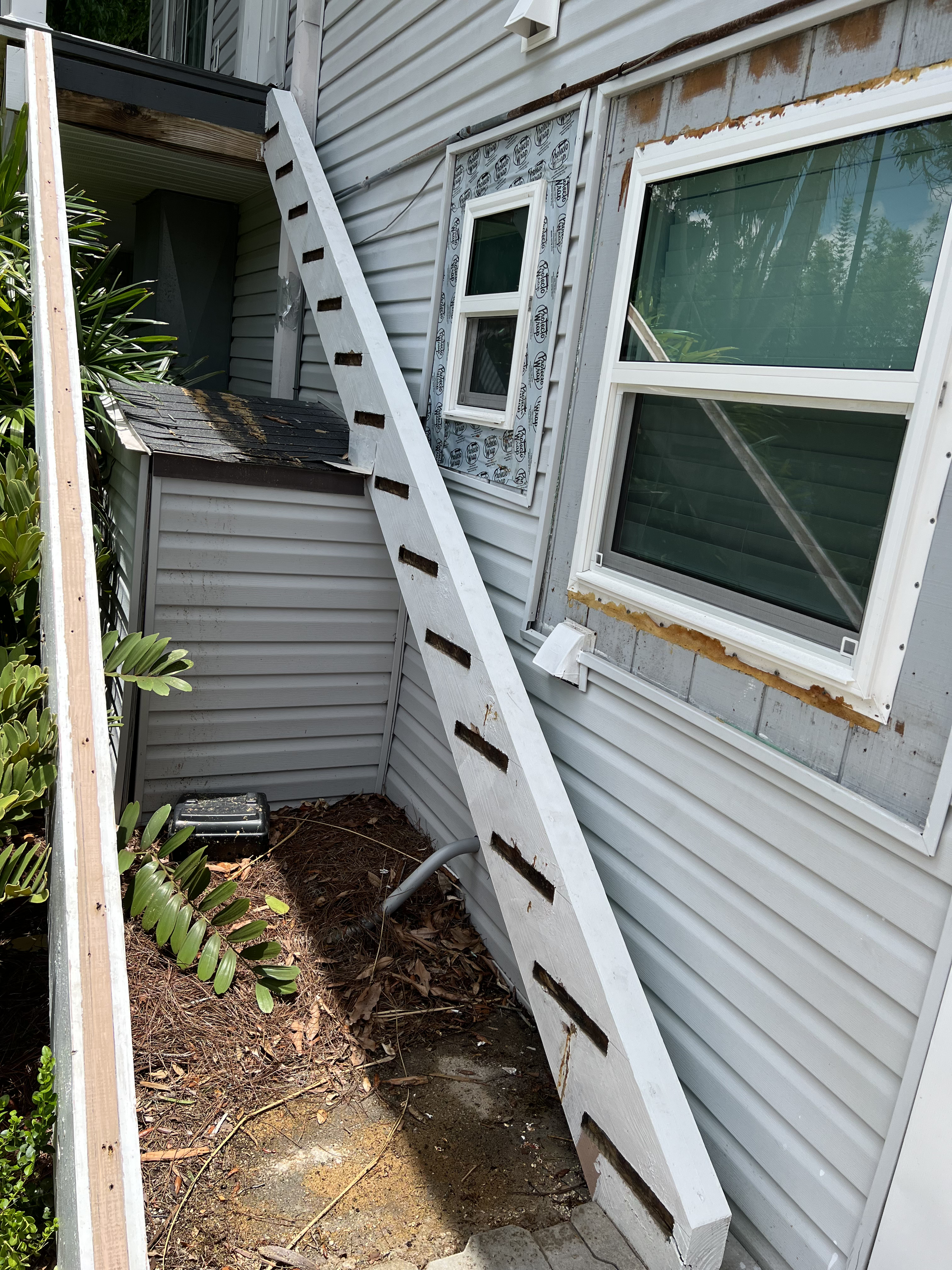 Exterior view of a house with siding, showing a partially removed window and an old window frame, with some construction materials like a ladder and debris on the ground.