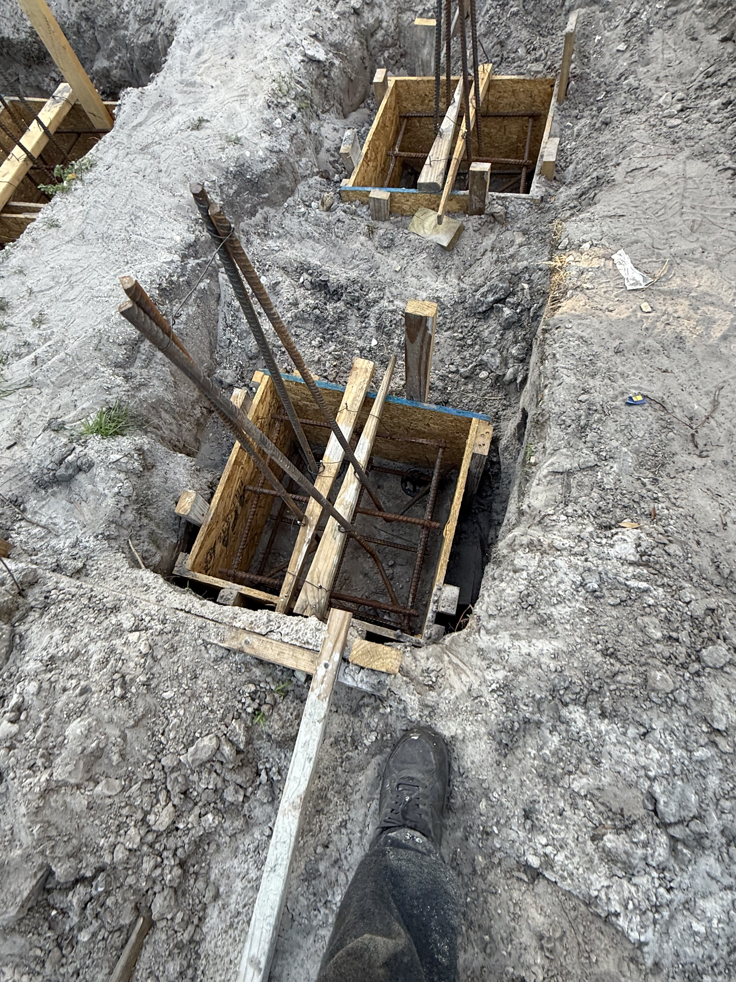 Construction site showing a narrow excavated trench with wooden formwork and rebar, and a person's muddy shoes in the foreground.