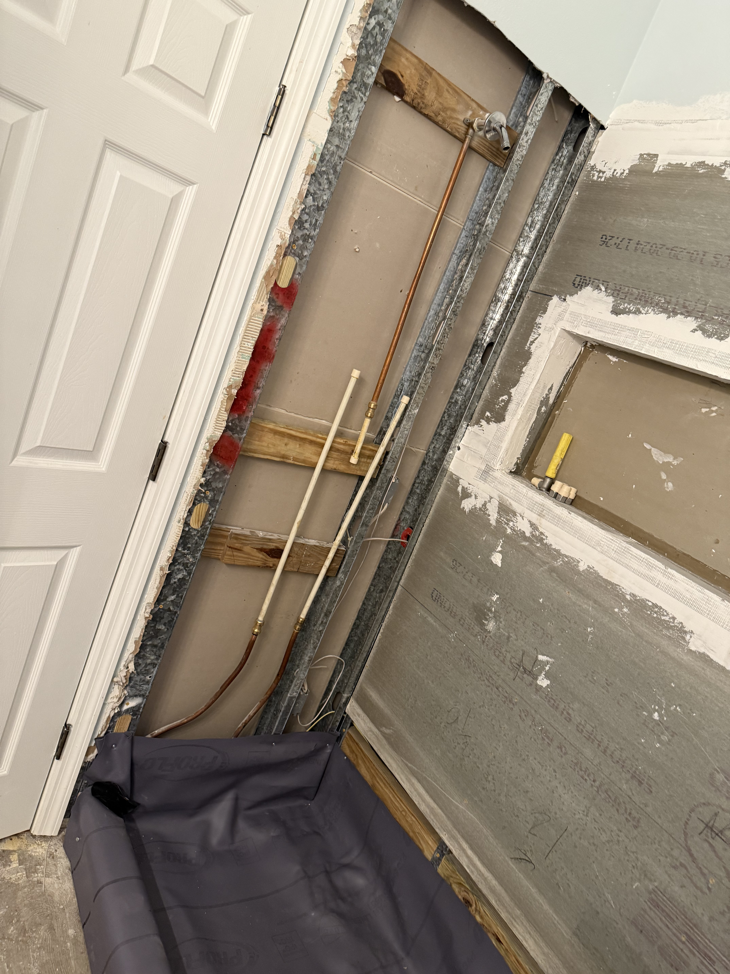 Construction or renovation site with exposed pipes, framing, and drywall. A white door is visible on the left, with a black bag or tarp at the bottom. The walls are partially finished with visible construction materials.