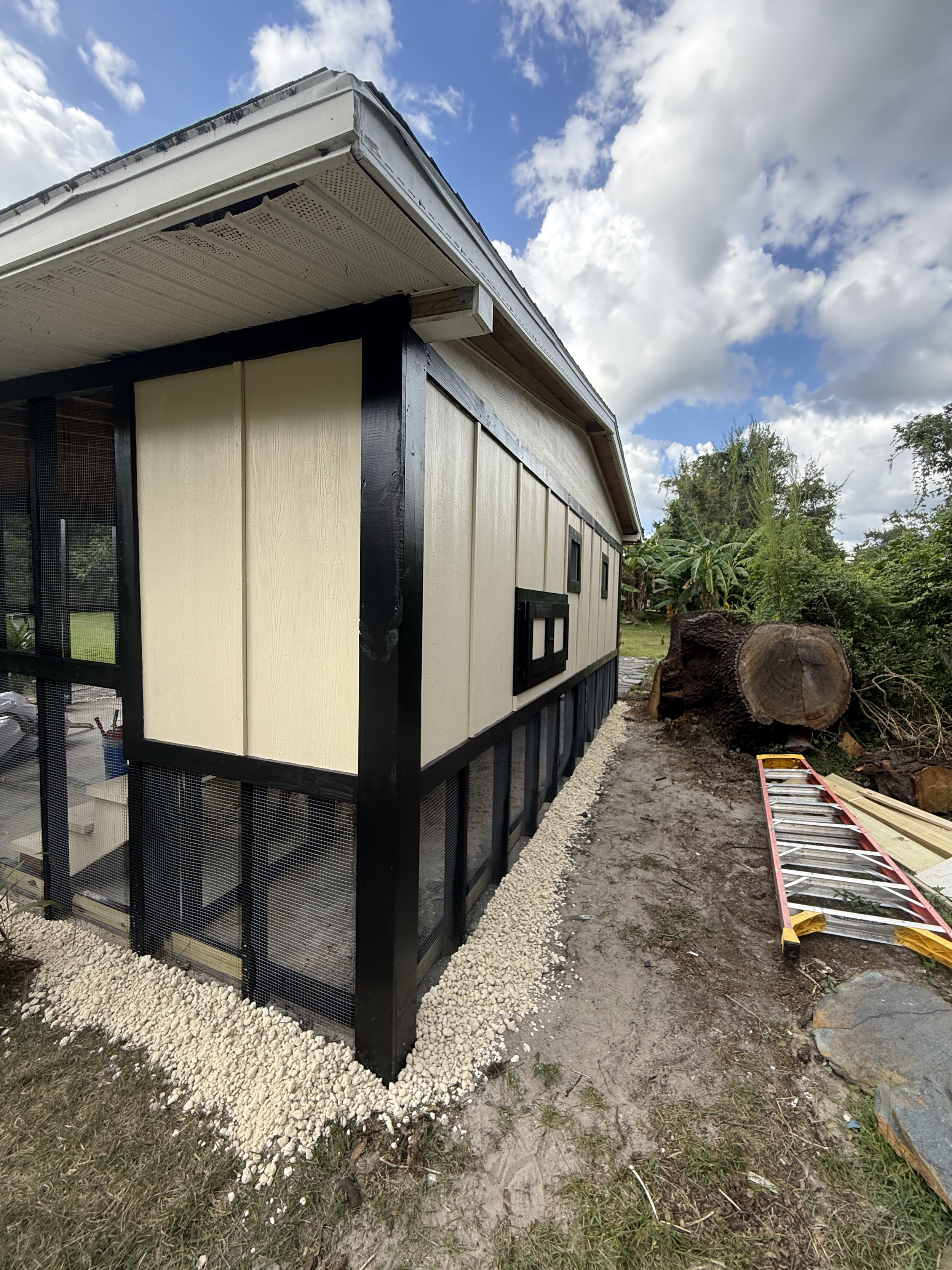 A small building or shed under construction with beige and black exterior walls, black framed windows, and a gravel foundation. The sky is partly cloudy with blue sky visible, and green trees and a large cut tree trunk are in the background. Construction materials and a ladder are on the ground near the building.
