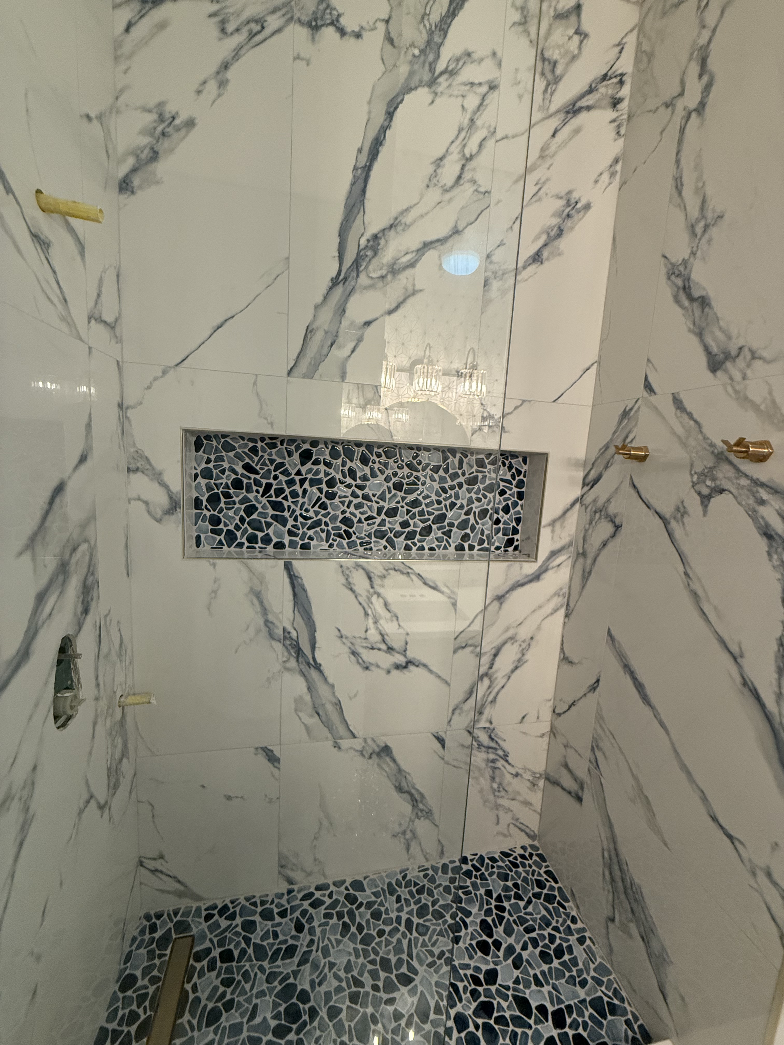 Bathroom with white marble walls featuring dark gray veining and mosaic tile flooring. A built-in niche with pebble mosaic tiles is visible on the wall. There are two gold-colored hooks on the wall, and a showerhead in the upper left corner.