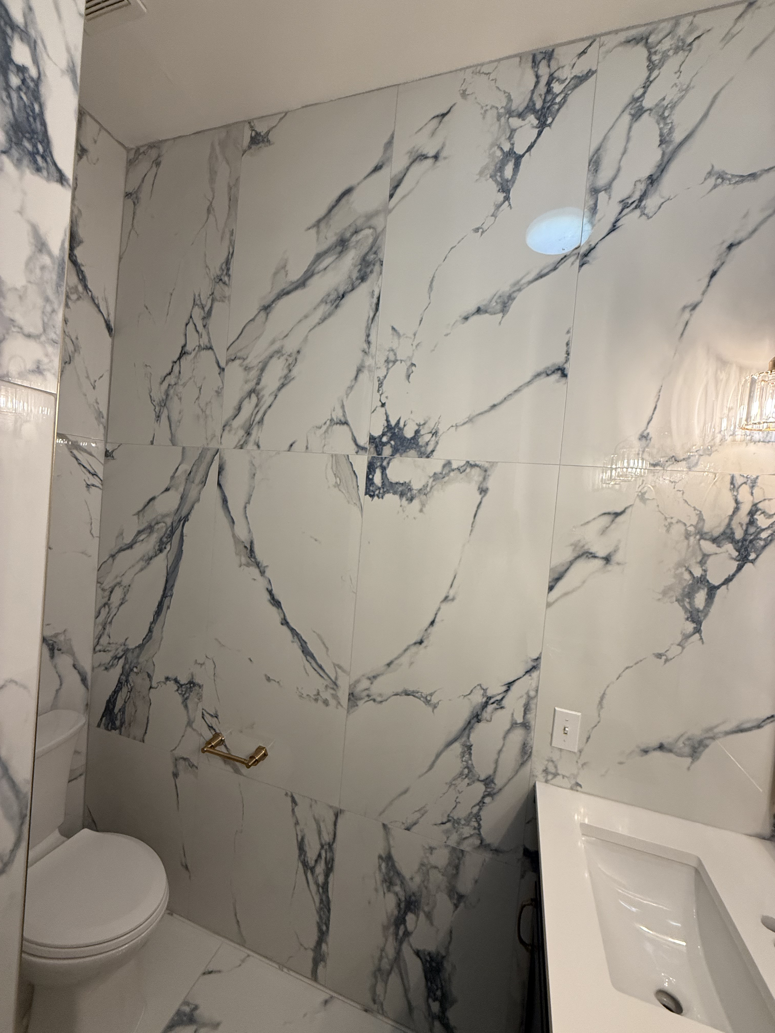 A bathroom with white marble walls and floor with black and gray veining. There is a white toilet on the left side and a black countertop with a white rectangular sink on the right side. A gold towel bar is mounted on the wall next to the toilet. An 