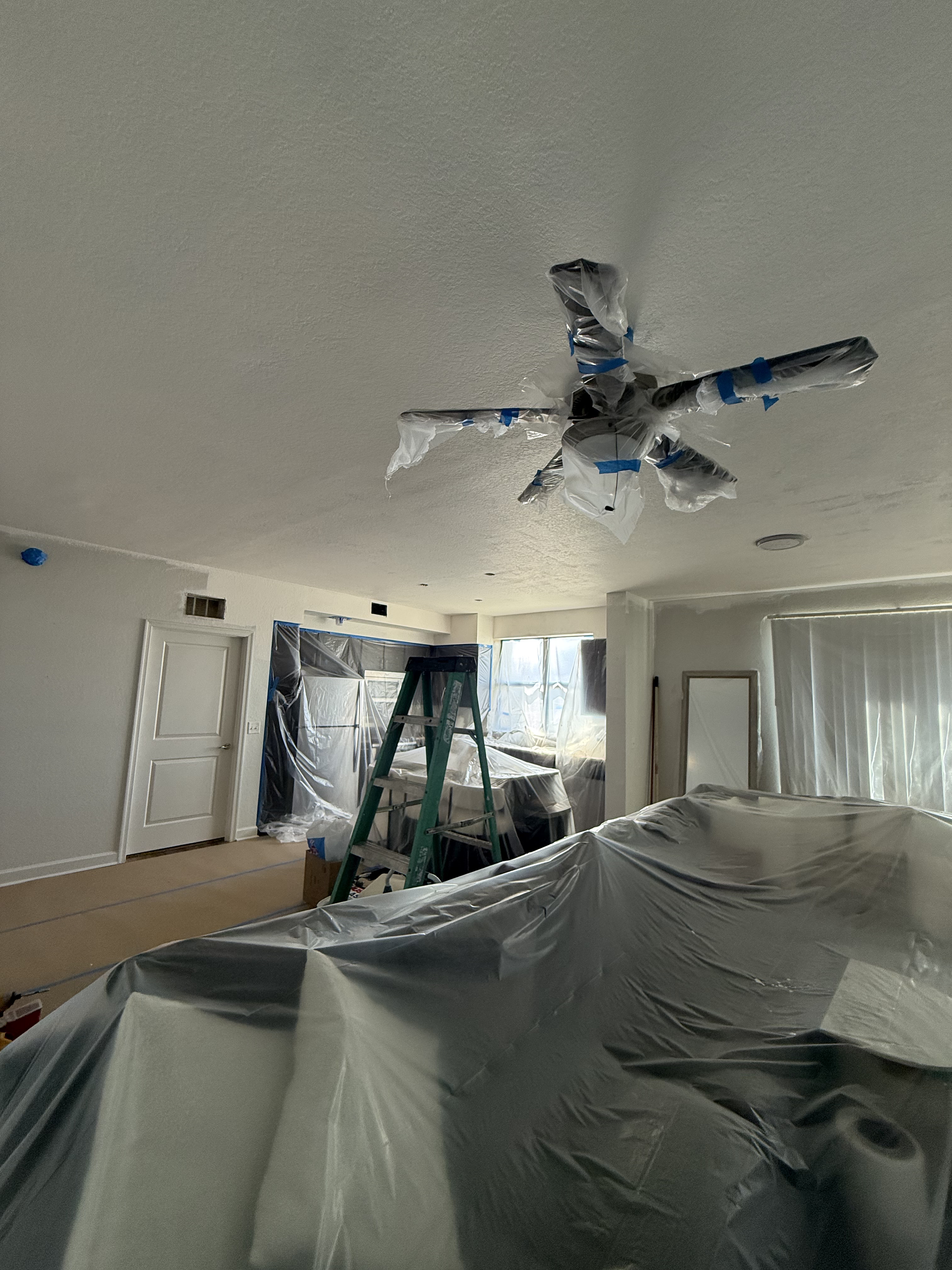 Living room undergoing renovation with plastic sheets covering furniture and a ladder, indicating painting or construction work in progress.