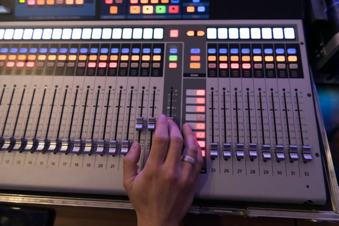 A person's left hand adjusting a knob on a professional audio mixing console with colorful faders, buttons, and sliders.