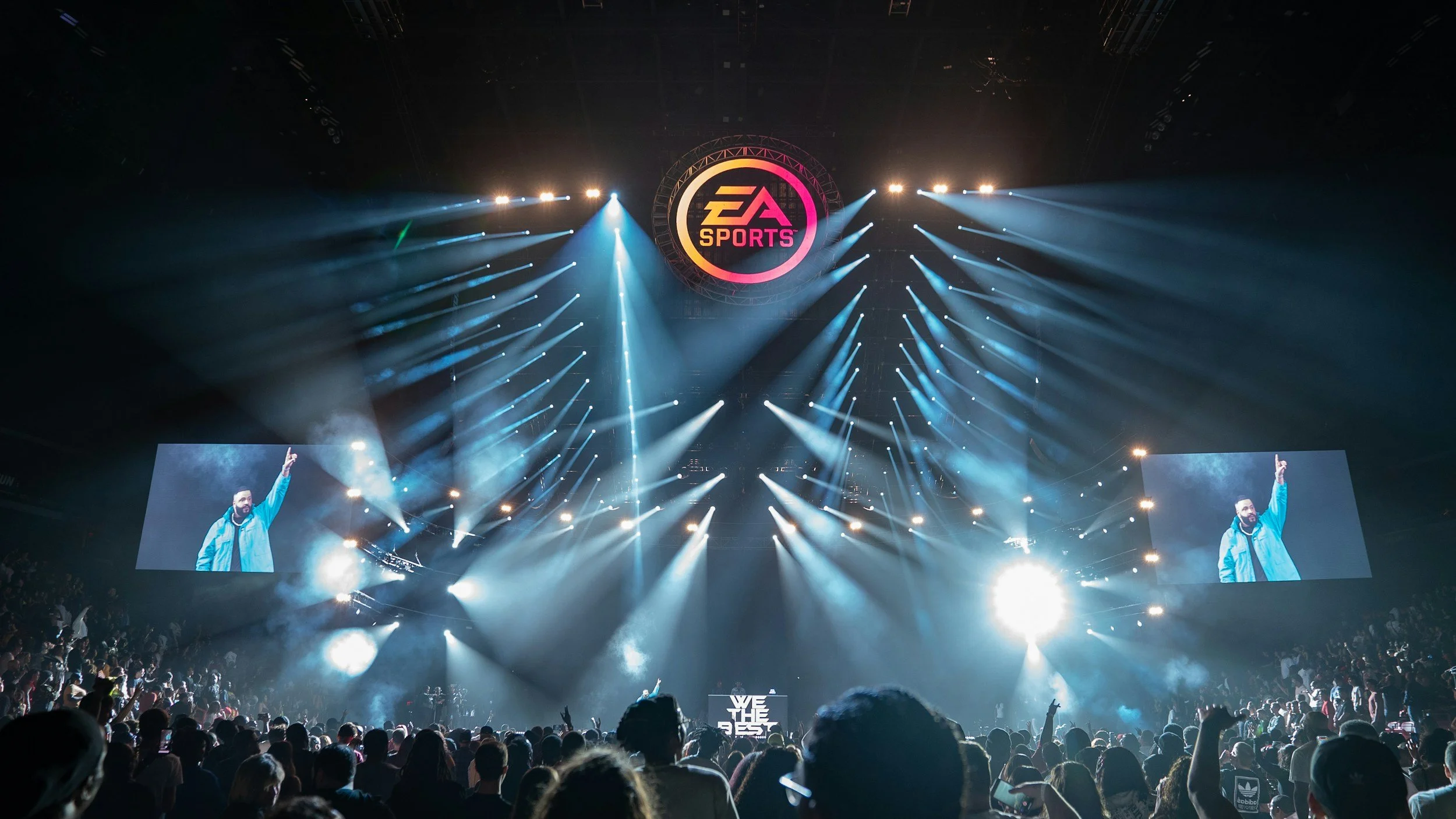 An esports event with a large crowd, stage lighting, and a screen displaying a person. The EA Sports logo is visible on a large circular display above the stage.