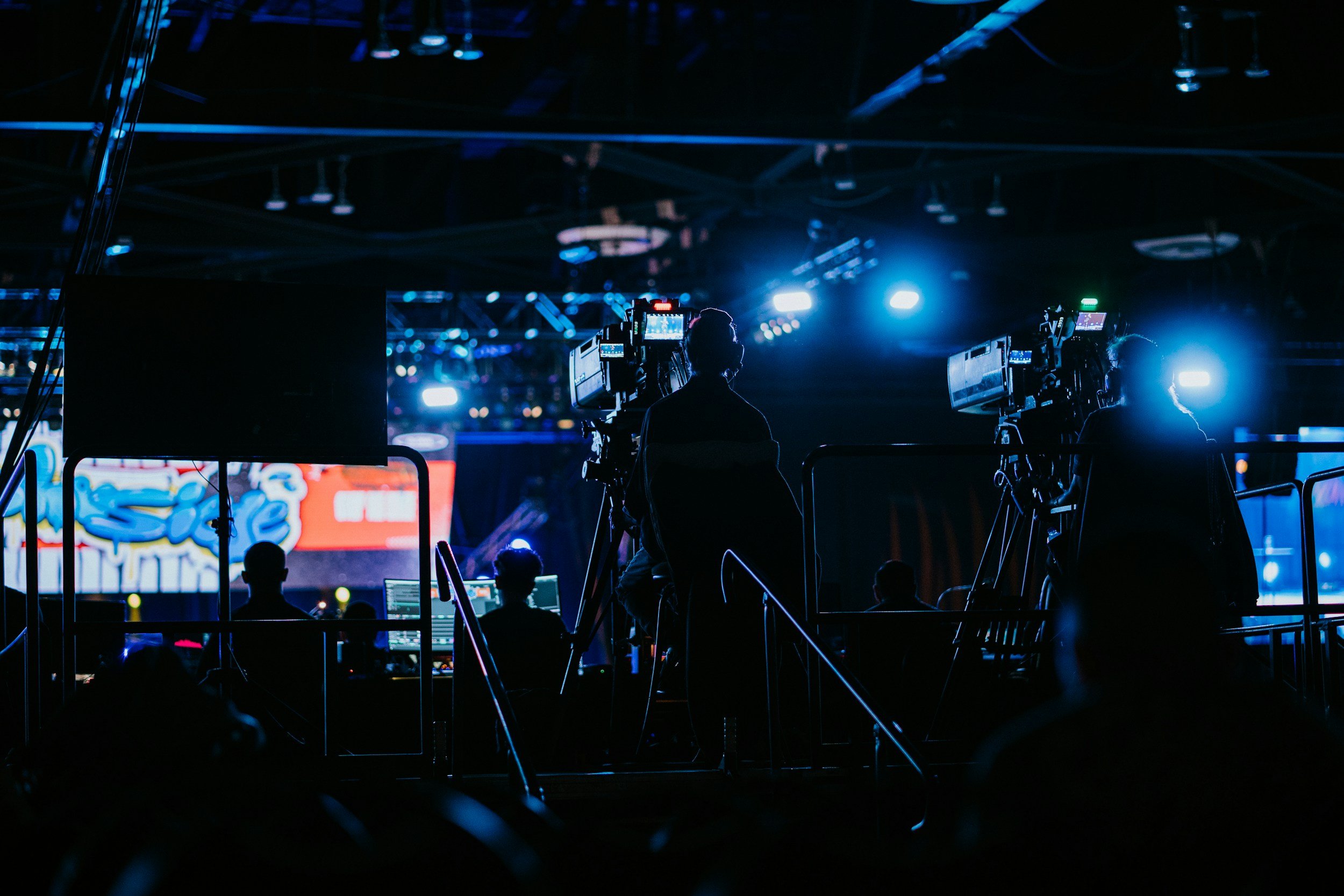 Behind the scenes of a live event production, multiple cameras and crew members are operating equipment with bright stage lights and digital screens visible in the background.