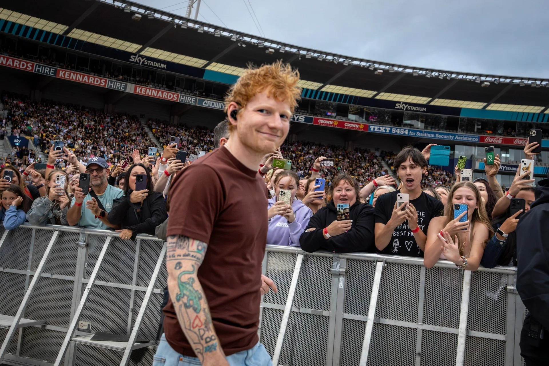 A young man with red hair and tattoos on his arm smiling at the camera, standing in front of a crowd at a stadium concert or event, with many people behind him taking photos and videos with their phones. The stadium has a large seating area with a roof and advertisements.