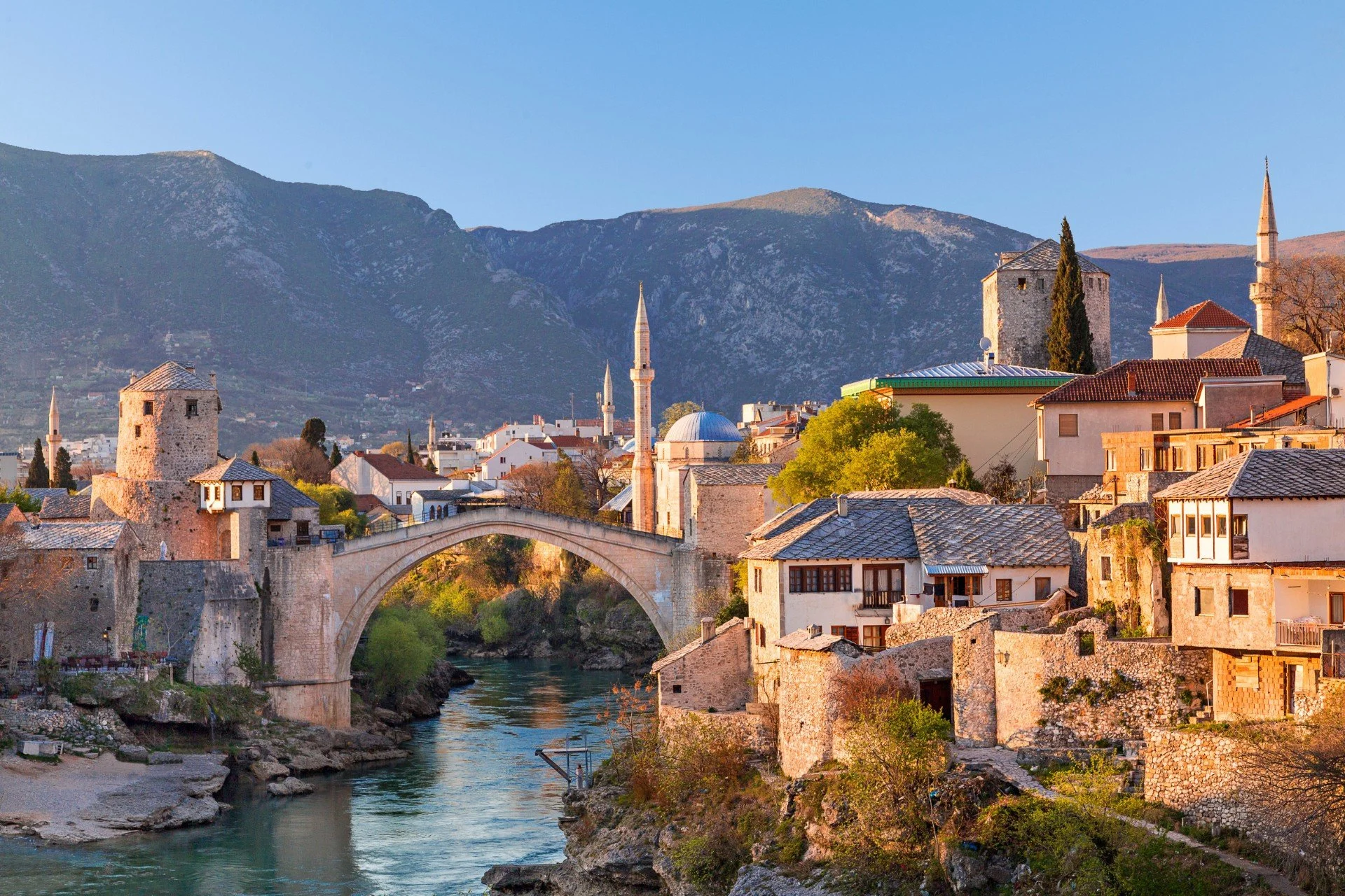 Historic town with stone buildings, mosques with minarets, a stone bridge over a river, and mountains in the background during daytime.
