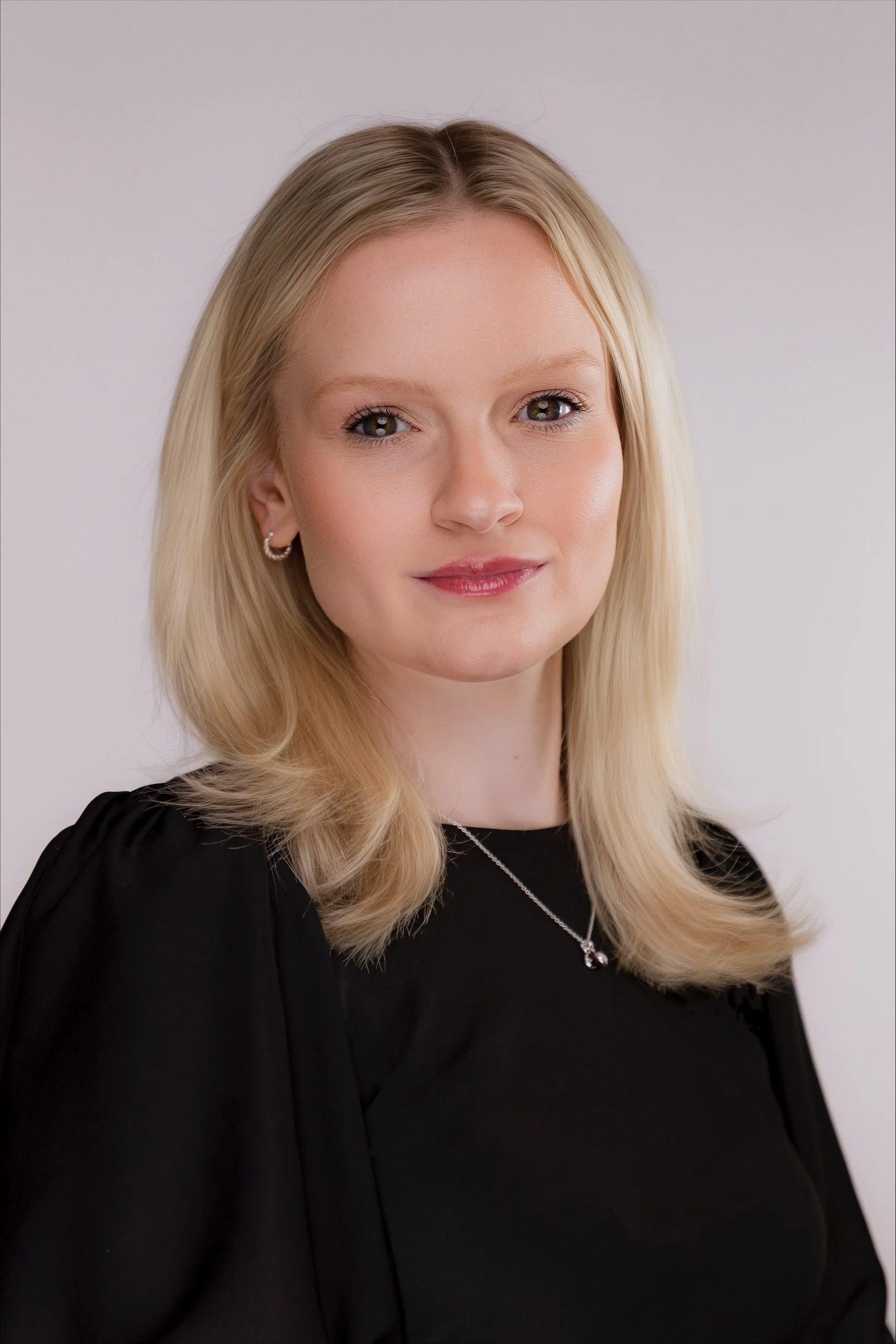 A woman with long blonde hair, wearing a black top, a silver necklace, and earrings, smiling at the camera against a plain light background.