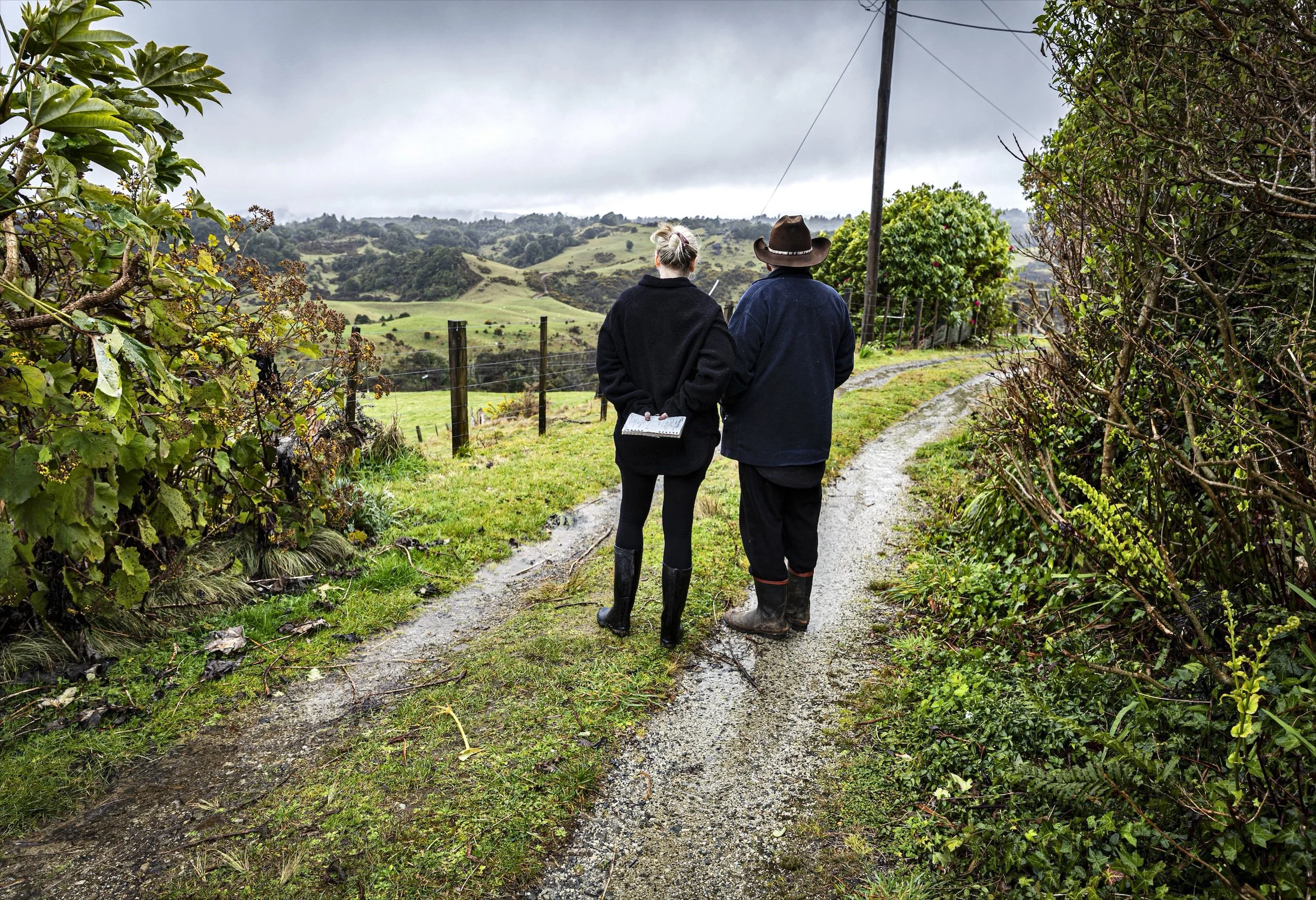 Two people walking along a dirt trail in a lush rural area with greenery and rolling hills, overcast sky, one with a notebook, the other with a cowboy hat.