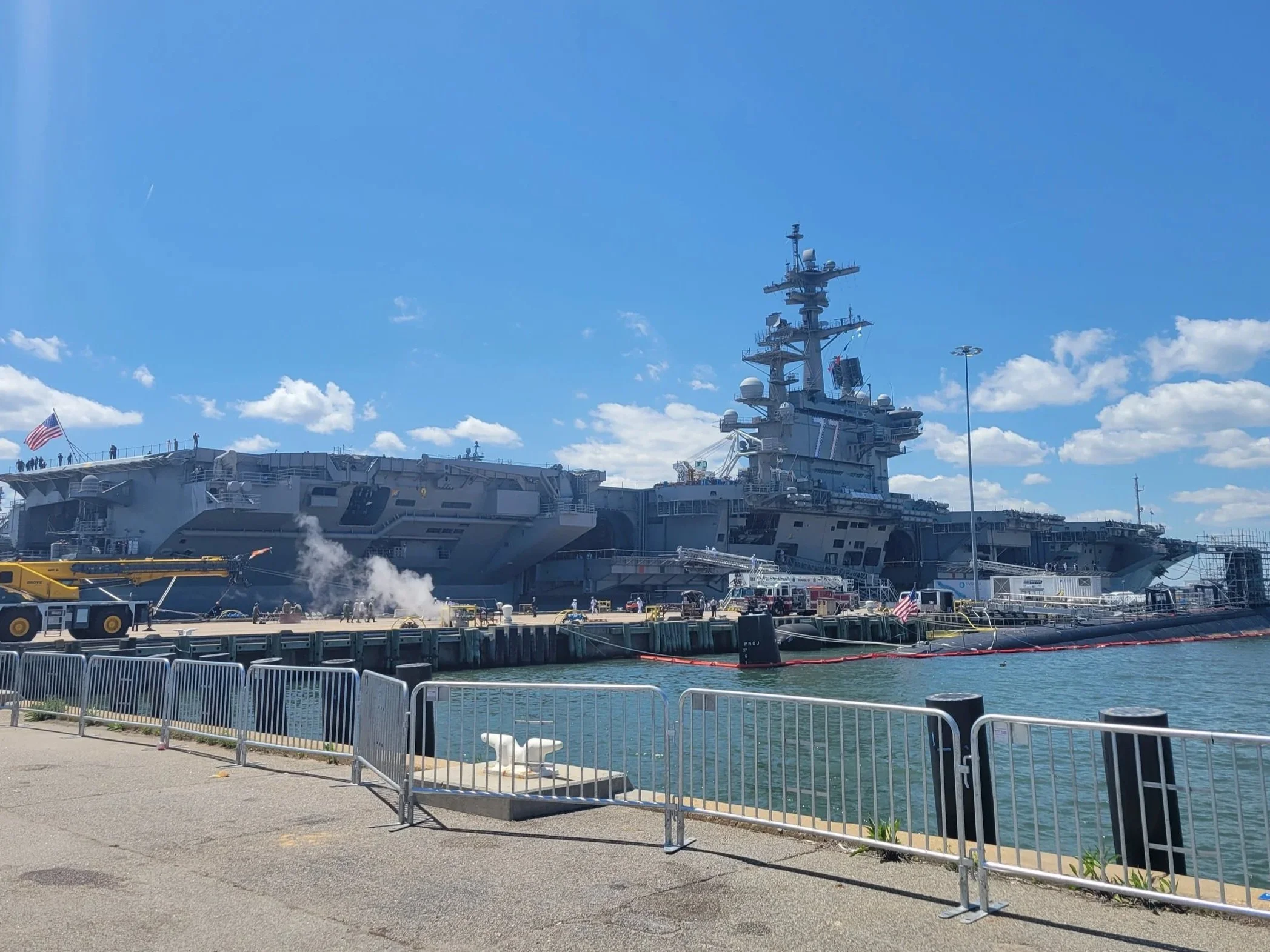 A large naval aircraft carrier docked at a pier under a blue sky with scattered clouds, surrounded by a metal barrier, with American flags flying on the ship and on the pier. USS George H.W. Bush was the ship the owner was stationed on.