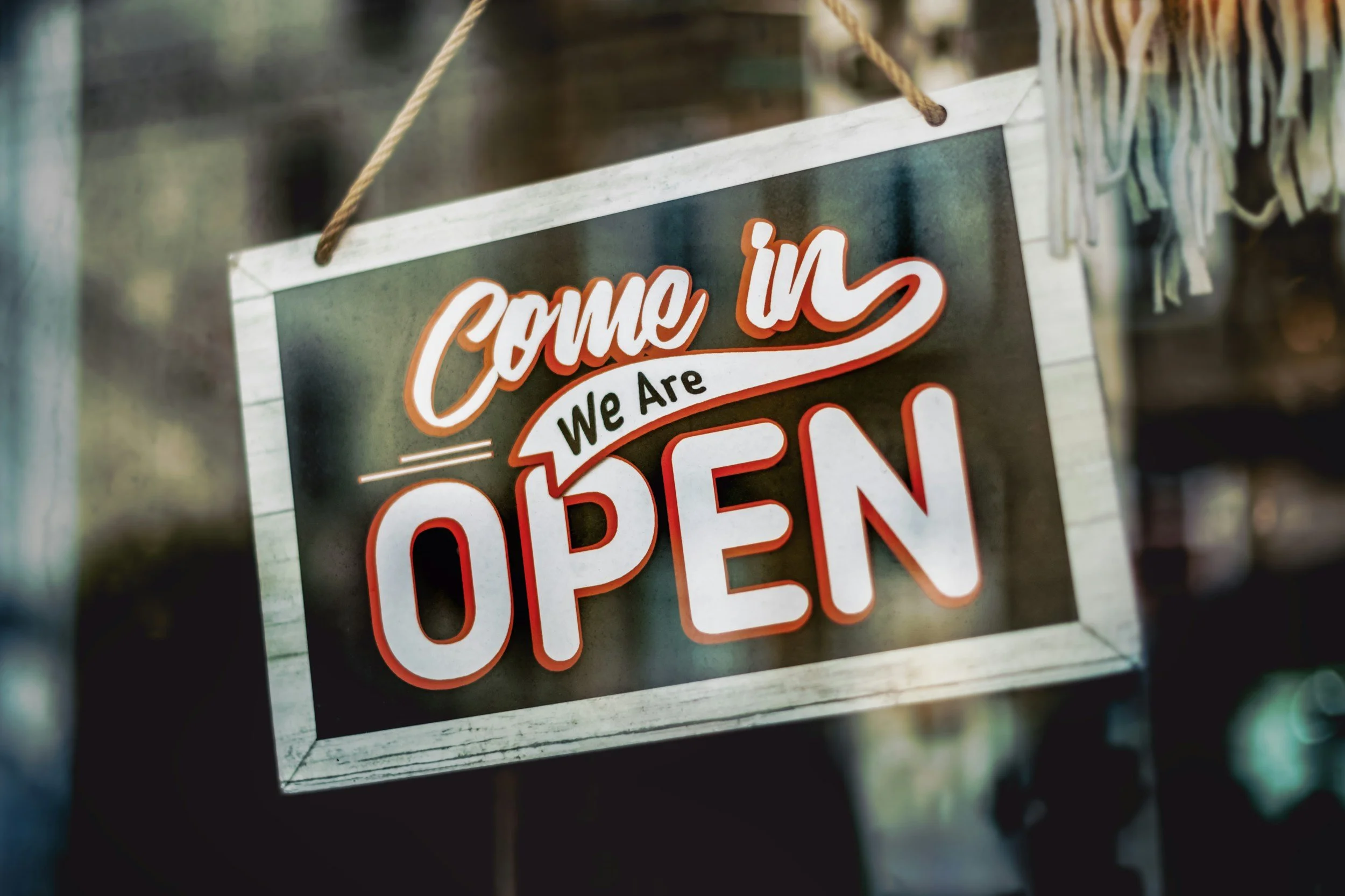 Sign hanging on a wall with the words "Come in, we are OPEN" in bright orange and white lettering.