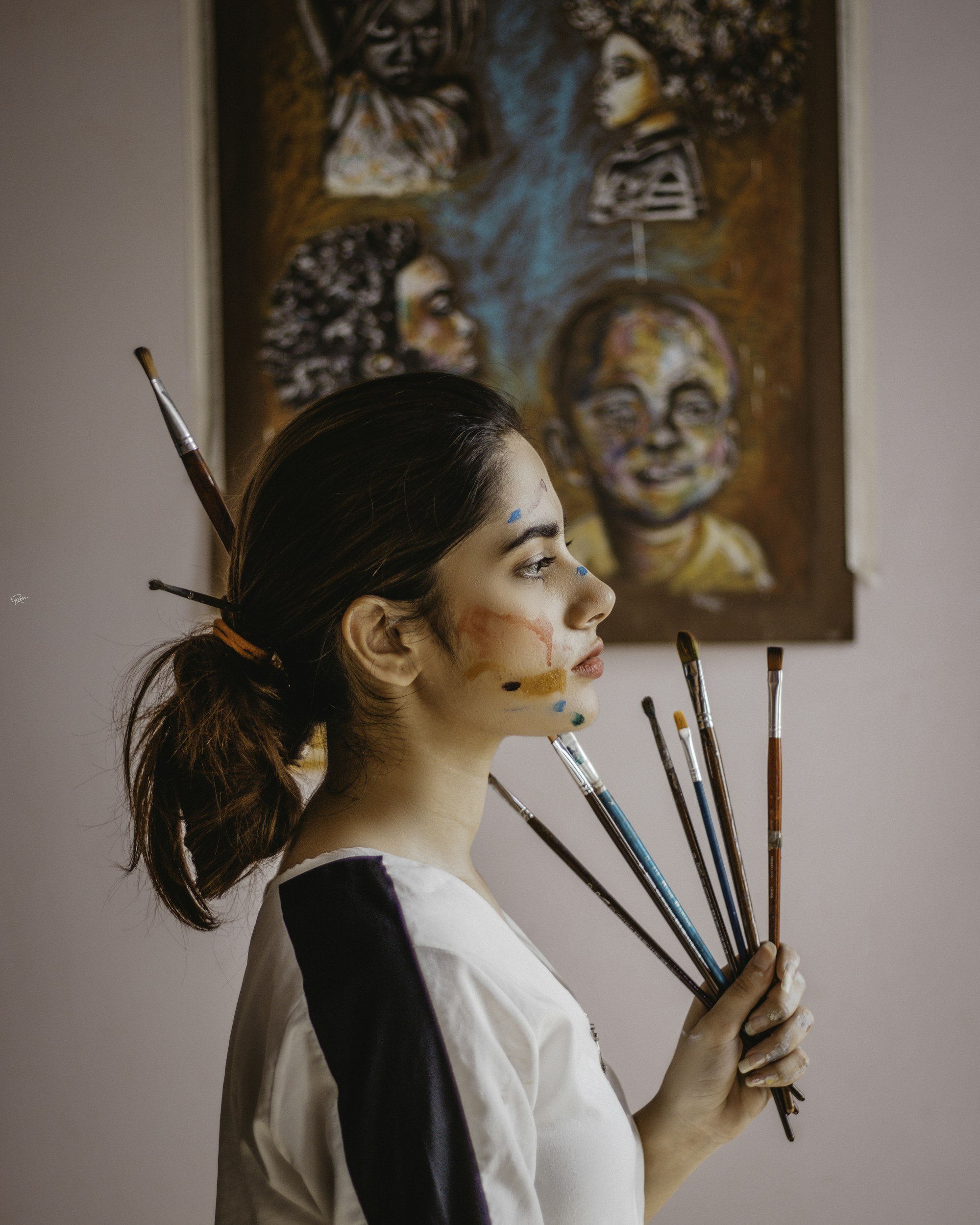 A woman with paint on her face and blue marks on her forehead and cheeks, holding several paintbrushes, standing in front of a colorful abstract portrait painting of various faces.