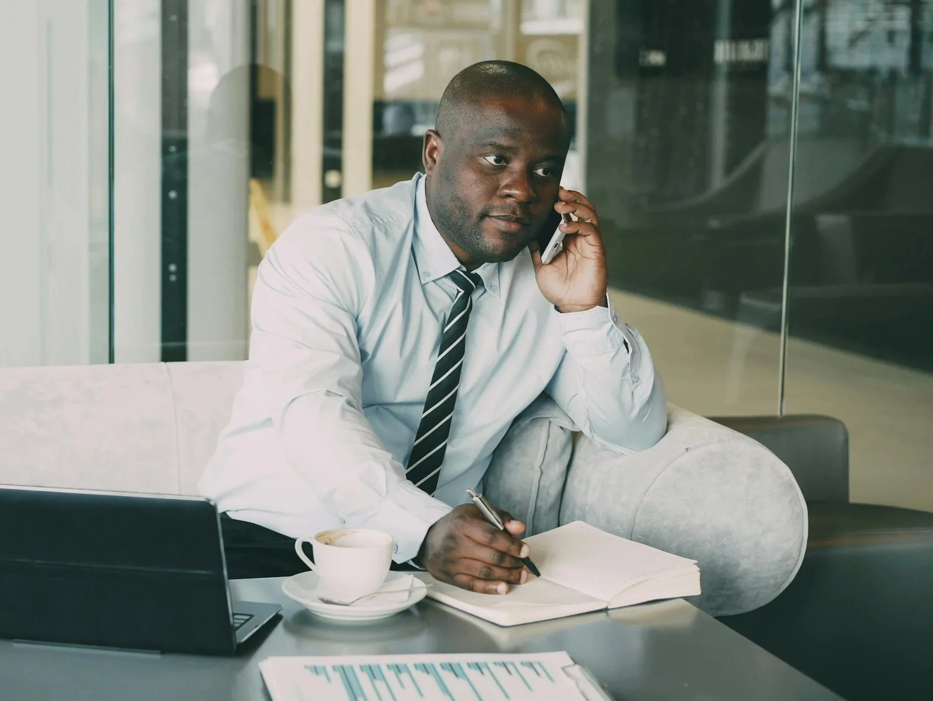 Man in dress shirt and tie talking on phone, sitting at desk with laptop, coffee cup, pen, notebook, and office documents.
