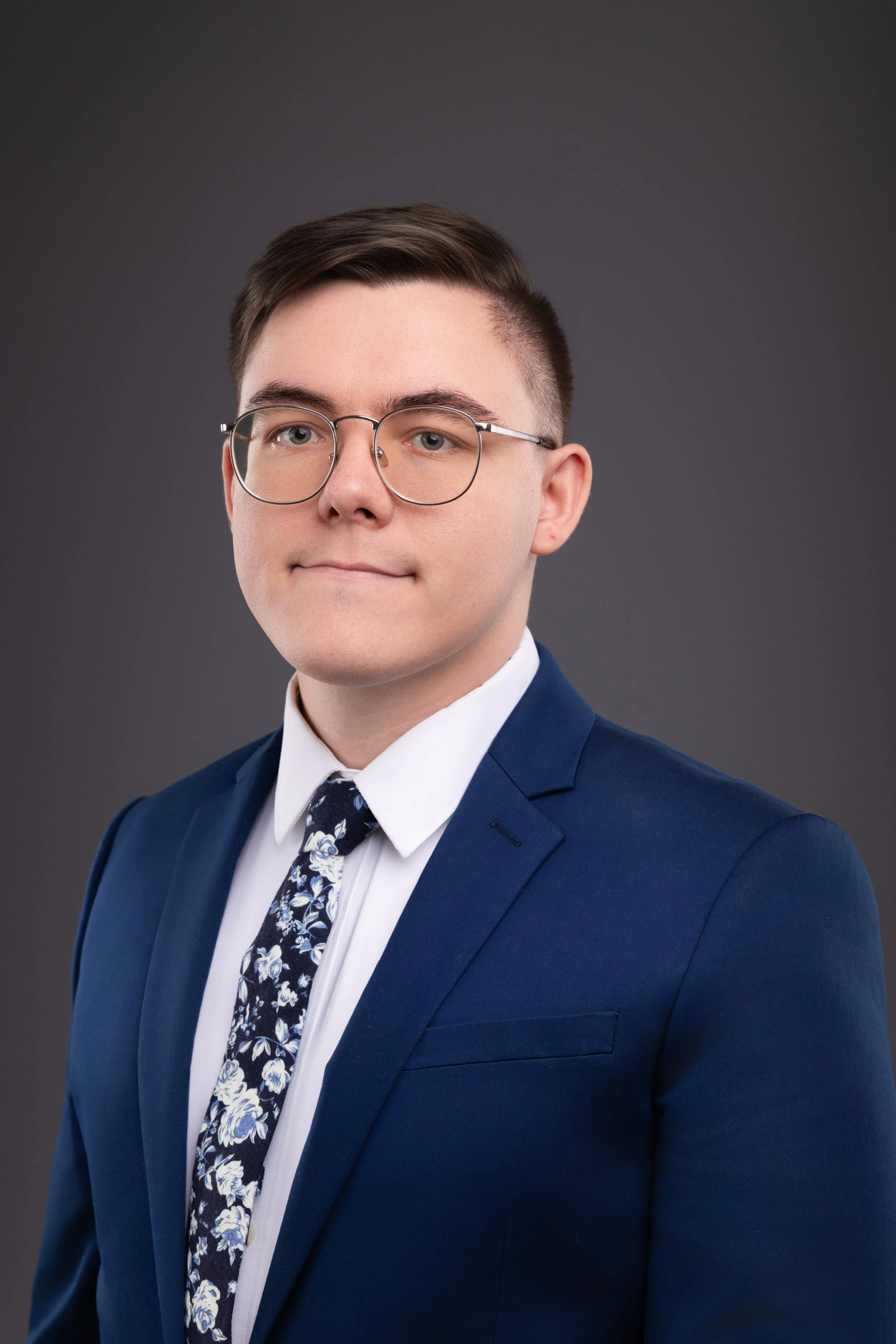 Professional portrait of the owner with glasses in a blue suit, white shirt, and floral tie against a gray background.