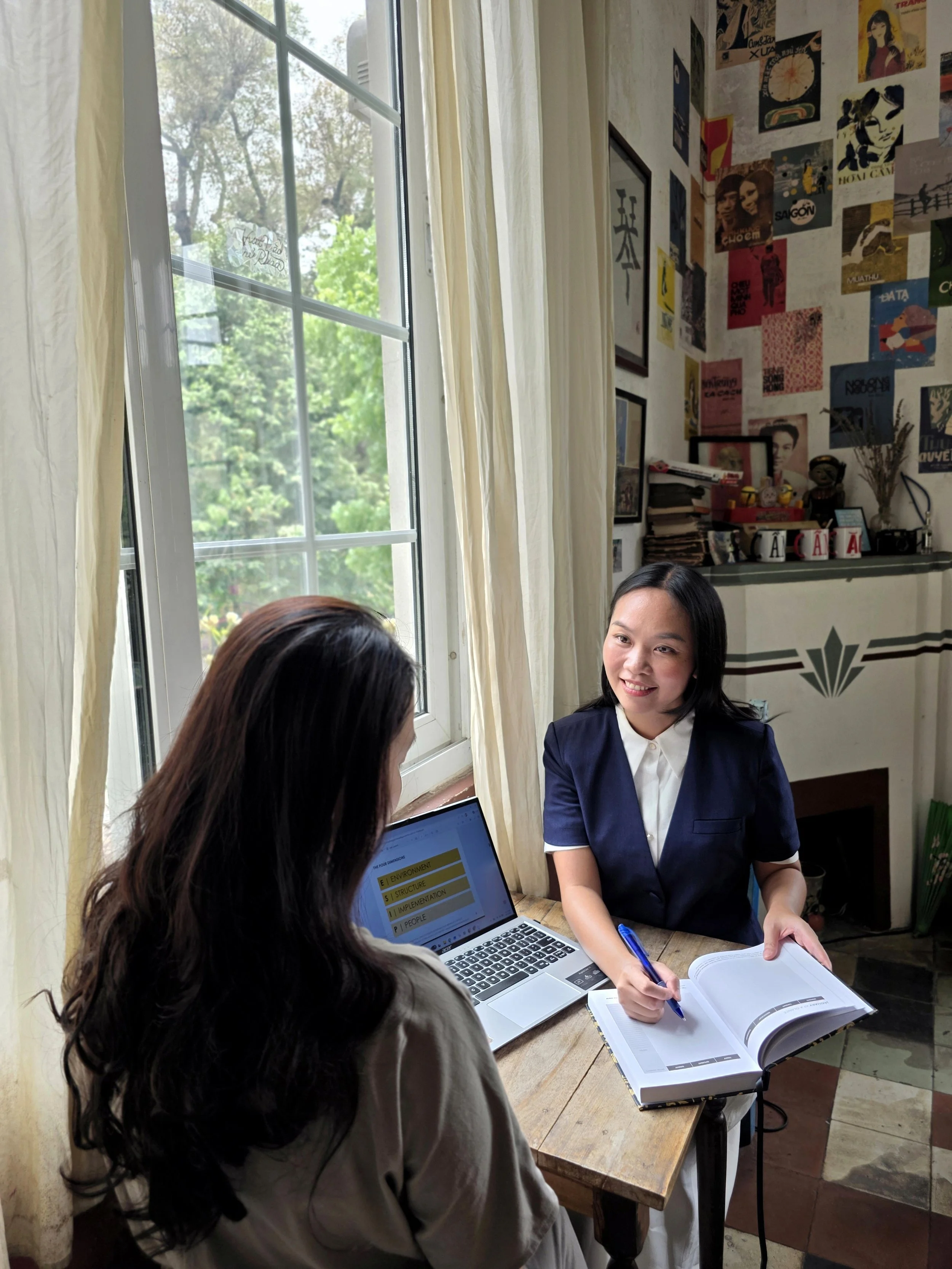 Two women having a discussion at a wooden table near a window. One woman with dark hair is facing away, while the other woman with shoulder-length black hair and a navy blazer is smiling, taking notes with a blue pen. The table has a laptop, notebook, and documents. The room has posters and pictures on the wall, a shelf with books, and a vase with dried flowers.