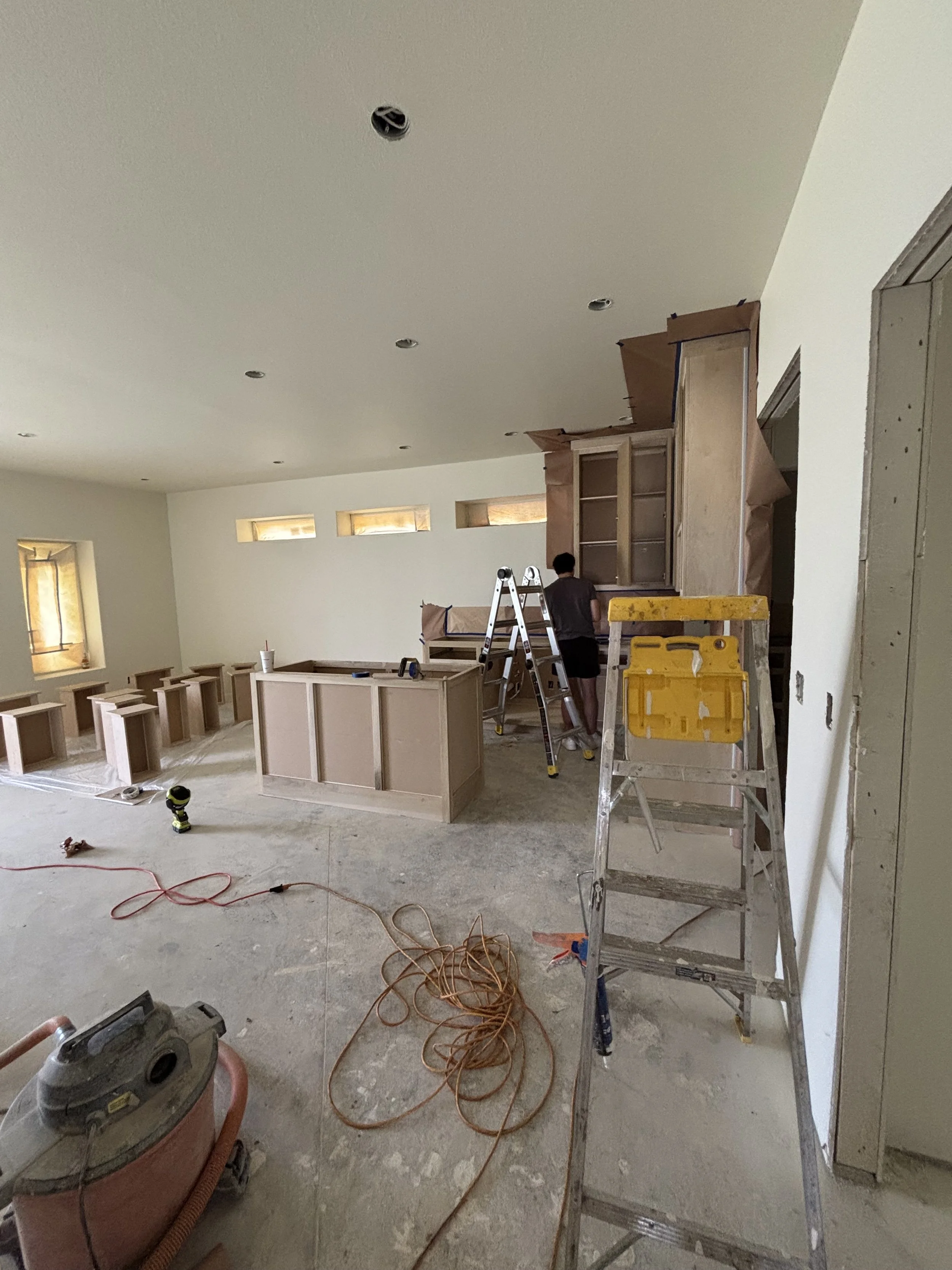 Interior of a house under renovation with construction tools, ladders, and workers installing cabinetry.