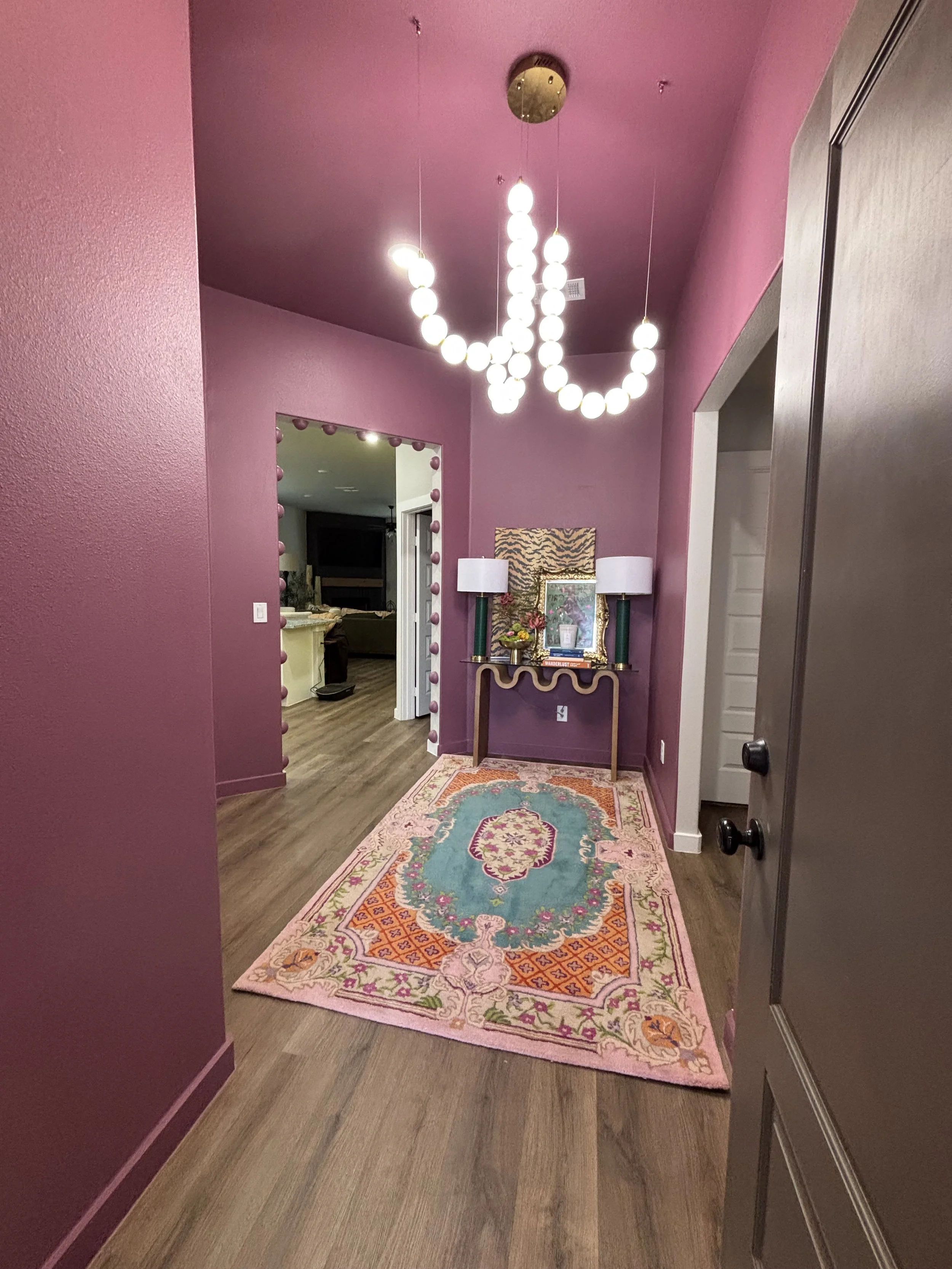 A hallway with purple walls, a colorful ornate rug, and a console table with lamps and artwork. A modern chandelier with multiple spherical bulbs hangs from the ceiling, and a doorway leads to another room.