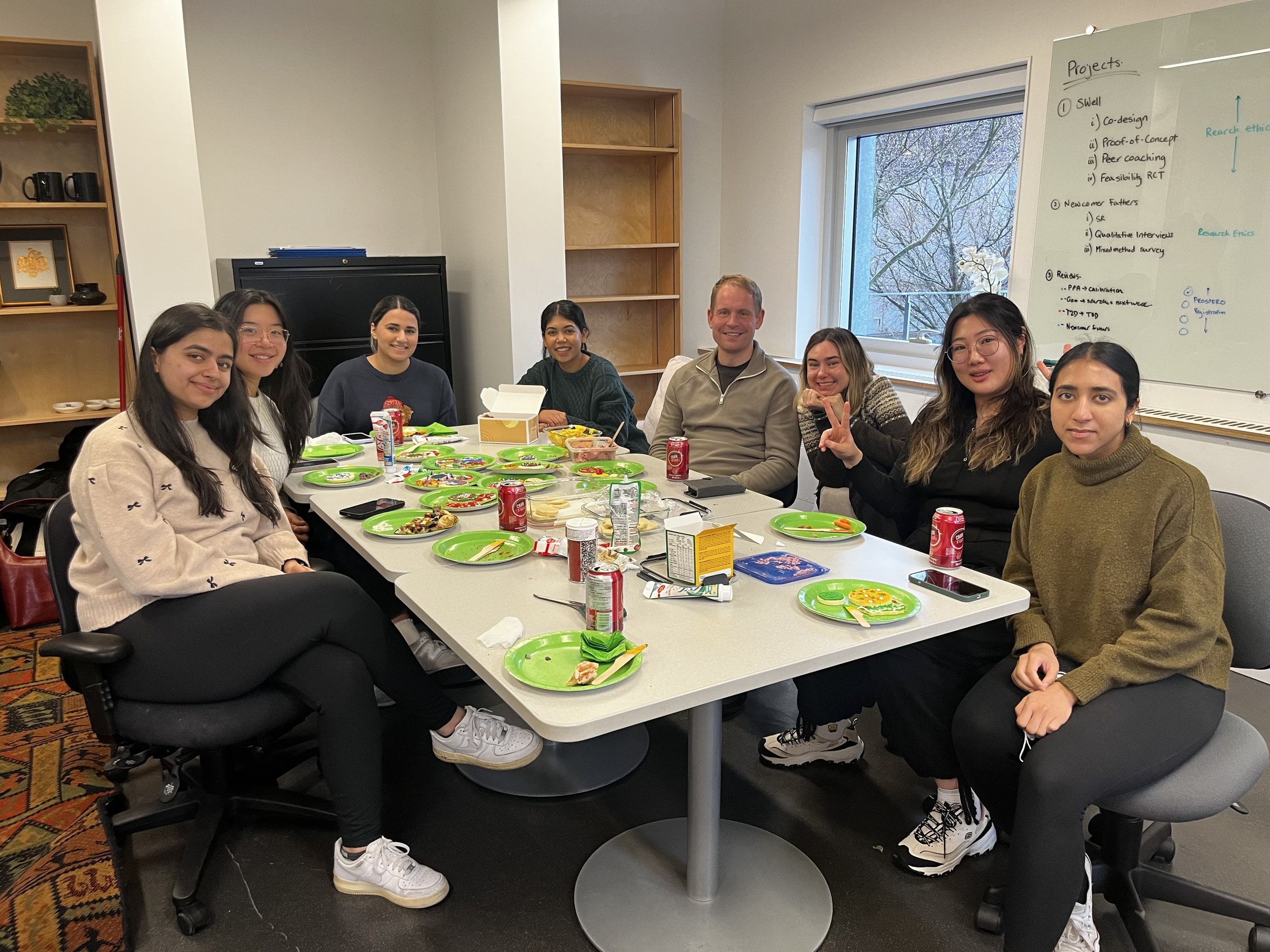A group of eight people sitting around a meeting table with green plates and food, smiling. Behind them are bookshelves, a whiteboard with notes, and a window showing trees outside.