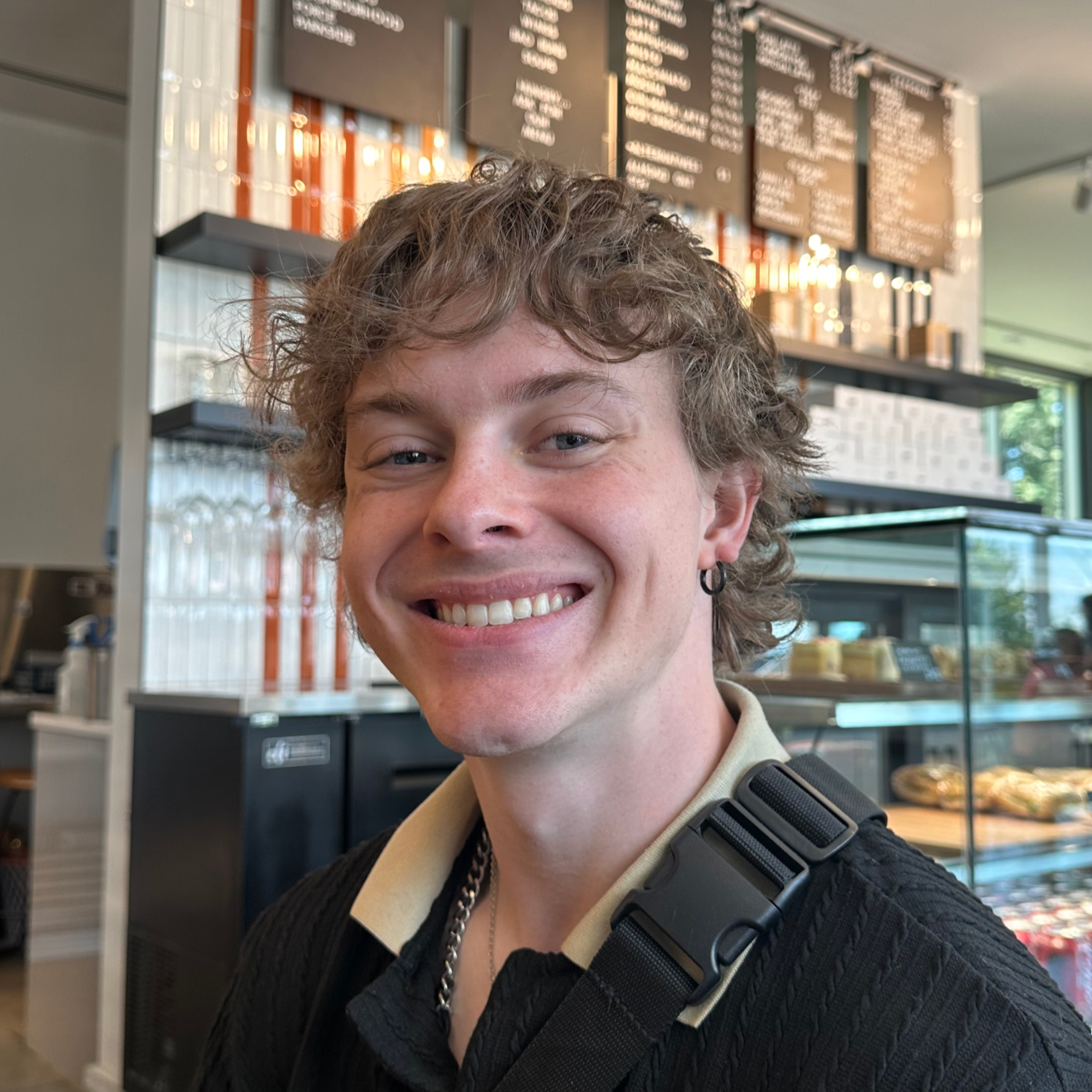 Close-up photo of a smiling young person with short curly hair, wearing a beige shirt, black sweater, silver chain necklace, and a black strap over their shoulder, inside a cafe or bakery with menu boards in the background.