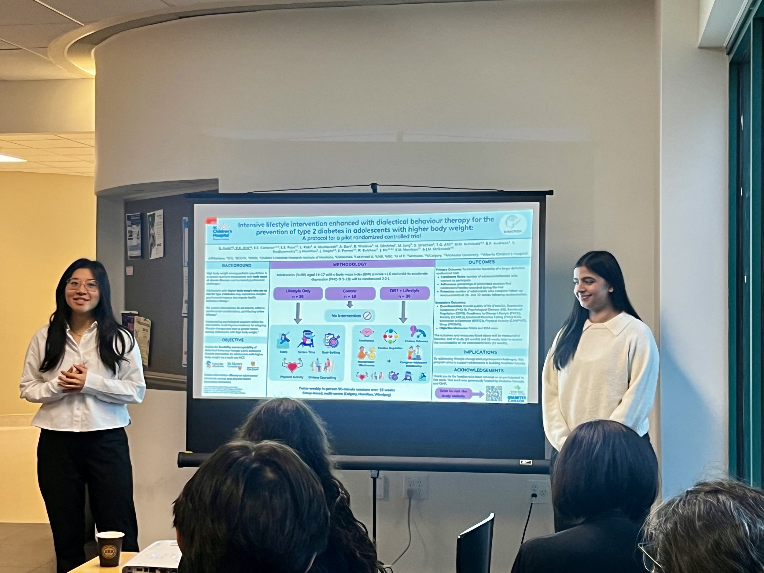 Two women standing next to a screen displaying a scientific research poster, with people seated in front of them attending a presentation in a conference room.
