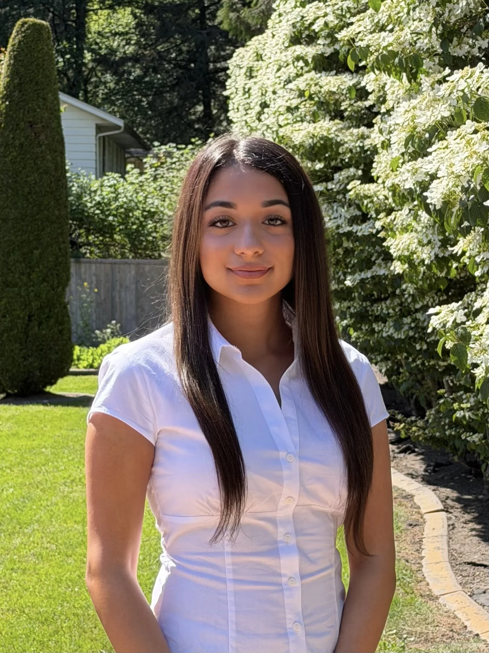 A young woman with long dark hair, wearing a white button-up shirt, standing outdoors in a garden with blooming white flowers and greenery, smiling softly.