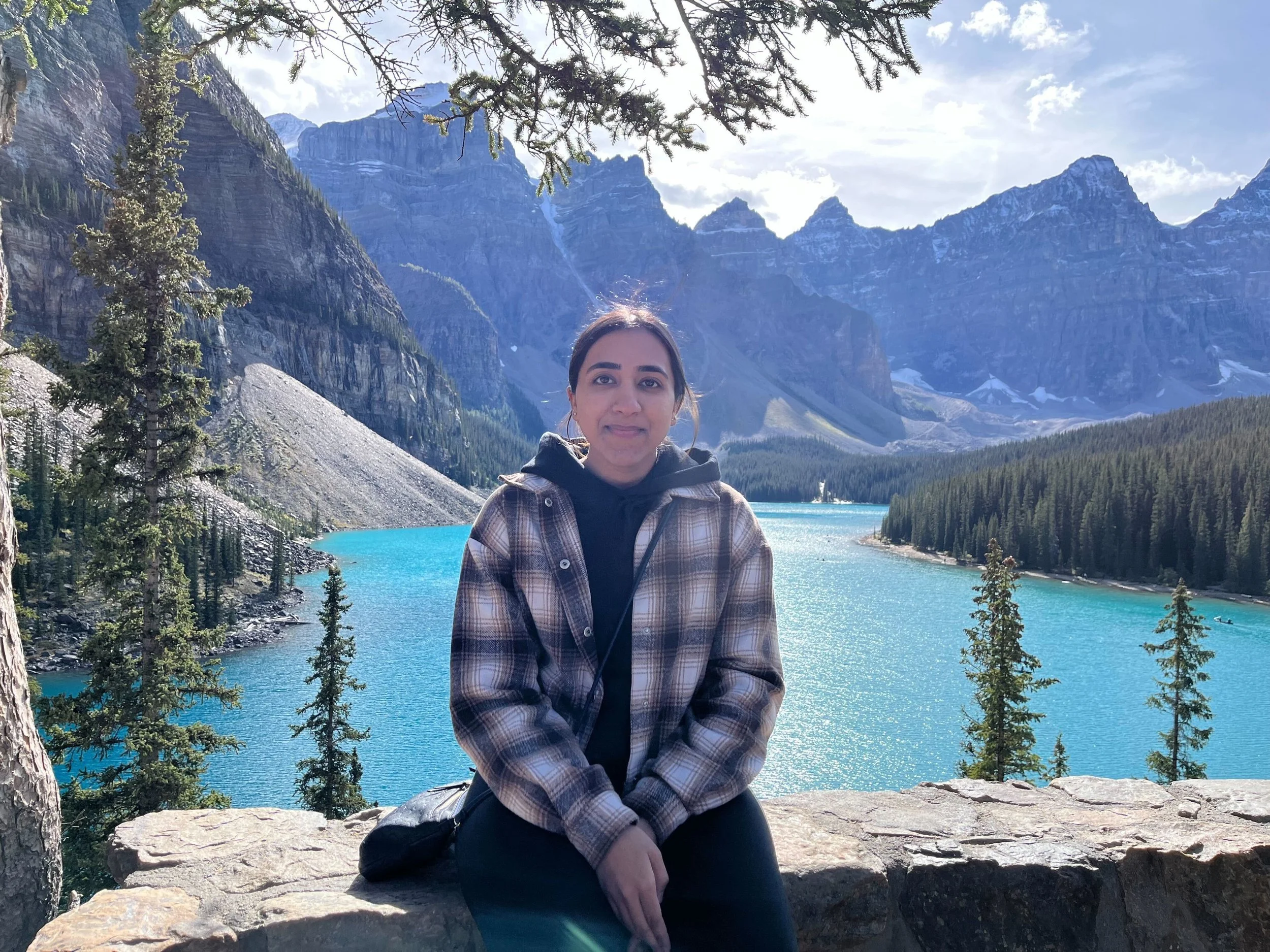 A woman sitting on a rock with a mountain lake behind her, surrounded by trees, in a scenic outdoor location.
