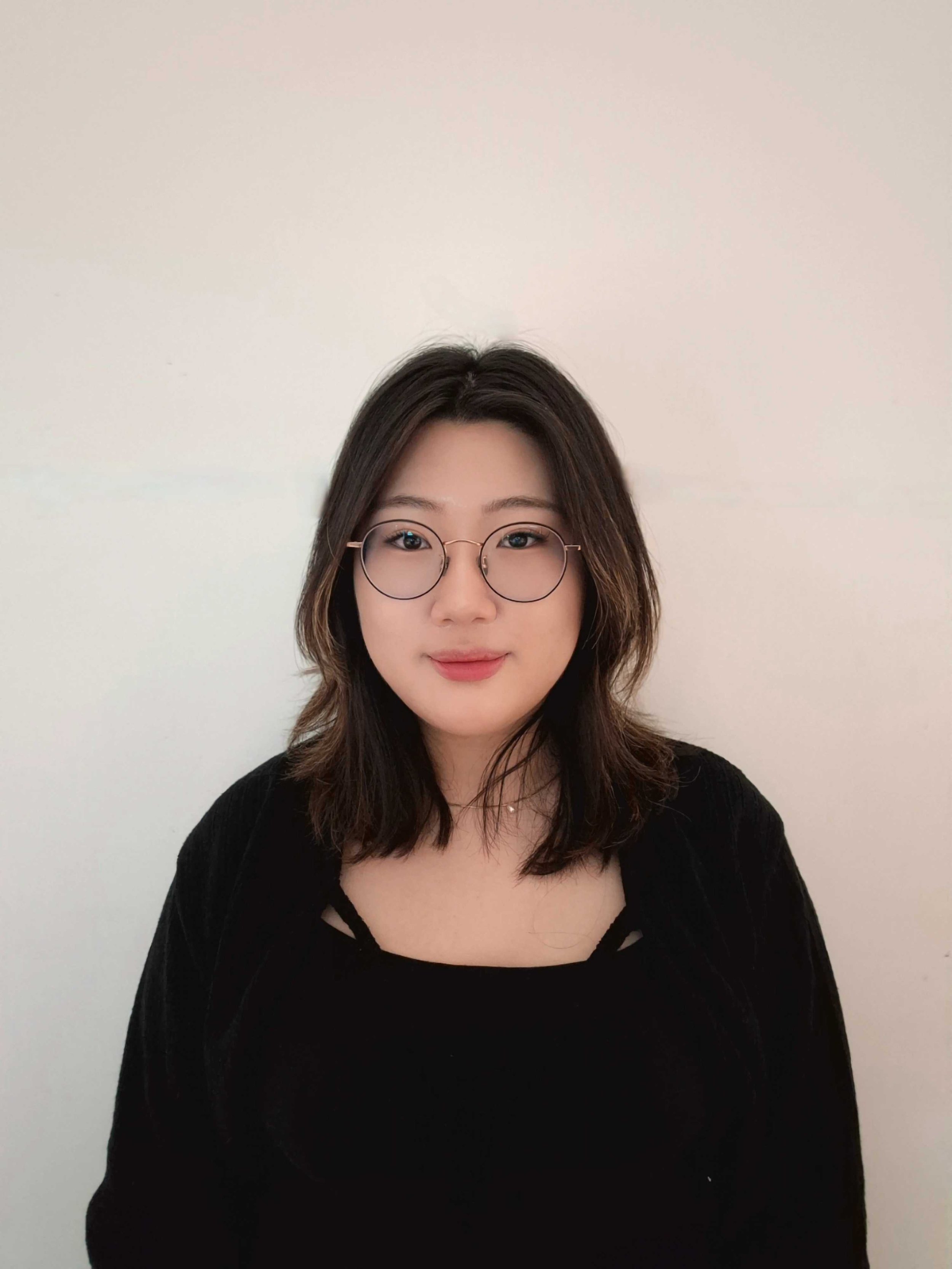 A young woman with shoulder-length dark hair, wearing glasses and a black top, is smiling softly in front of a plain white wall.