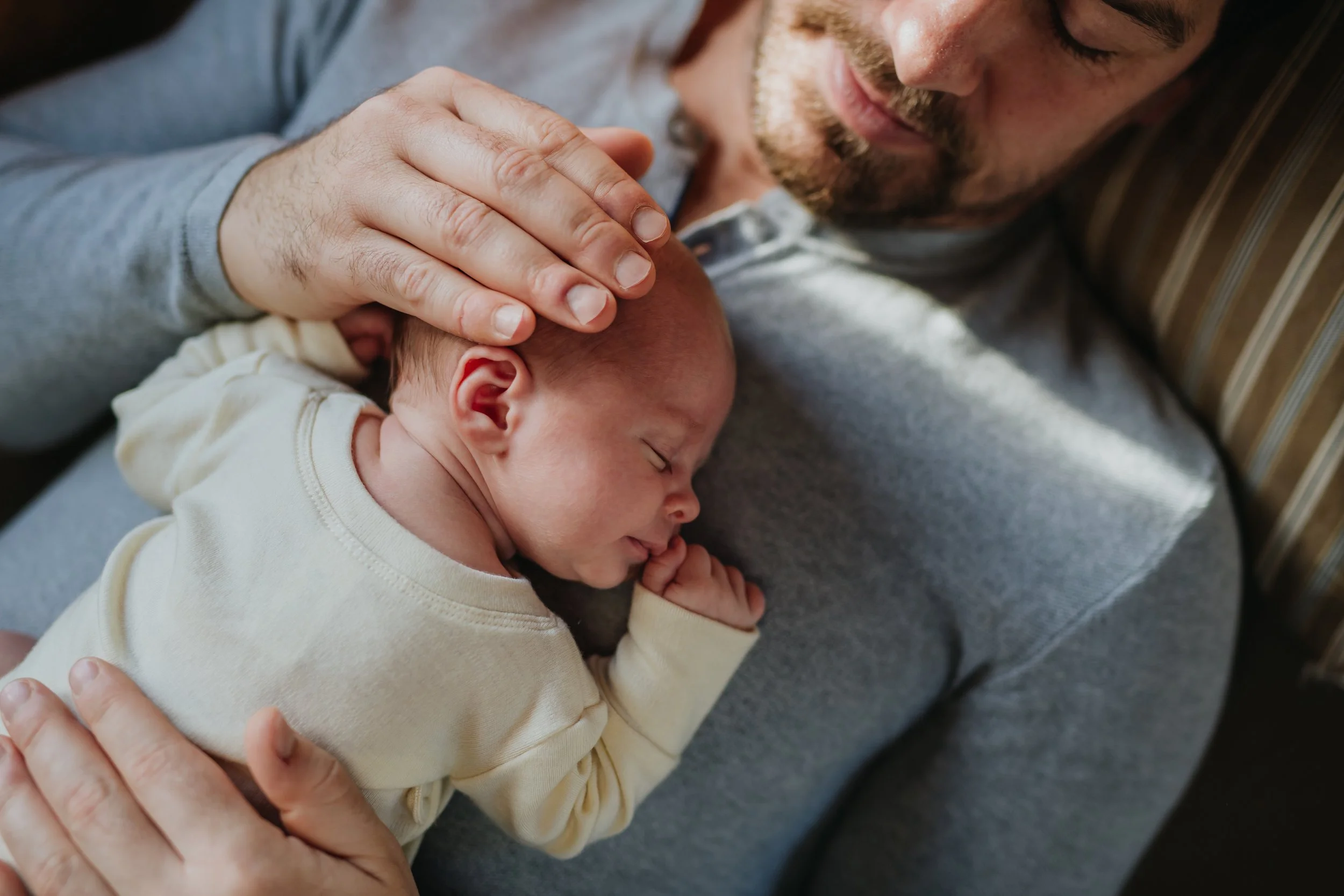 A close-up of a man holding a sleeping baby, resting on a gray couch. The man gently places his hand on the baby's head.