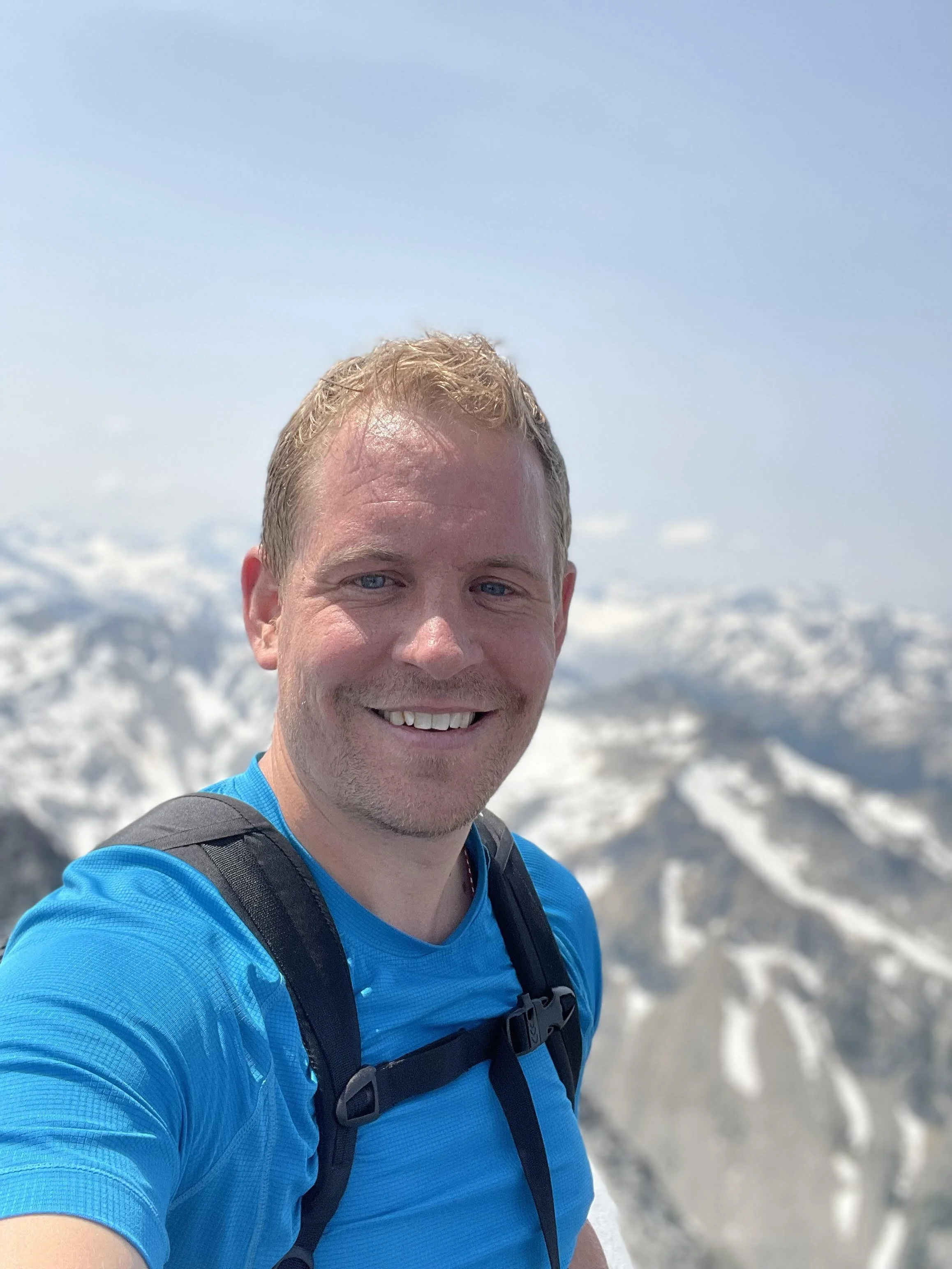 A smiling man with short blonde hair and blue eyes, wearing a blue shirt and a black backpack, standing outdoors on a snowy mountain with a clear blue sky in the background.