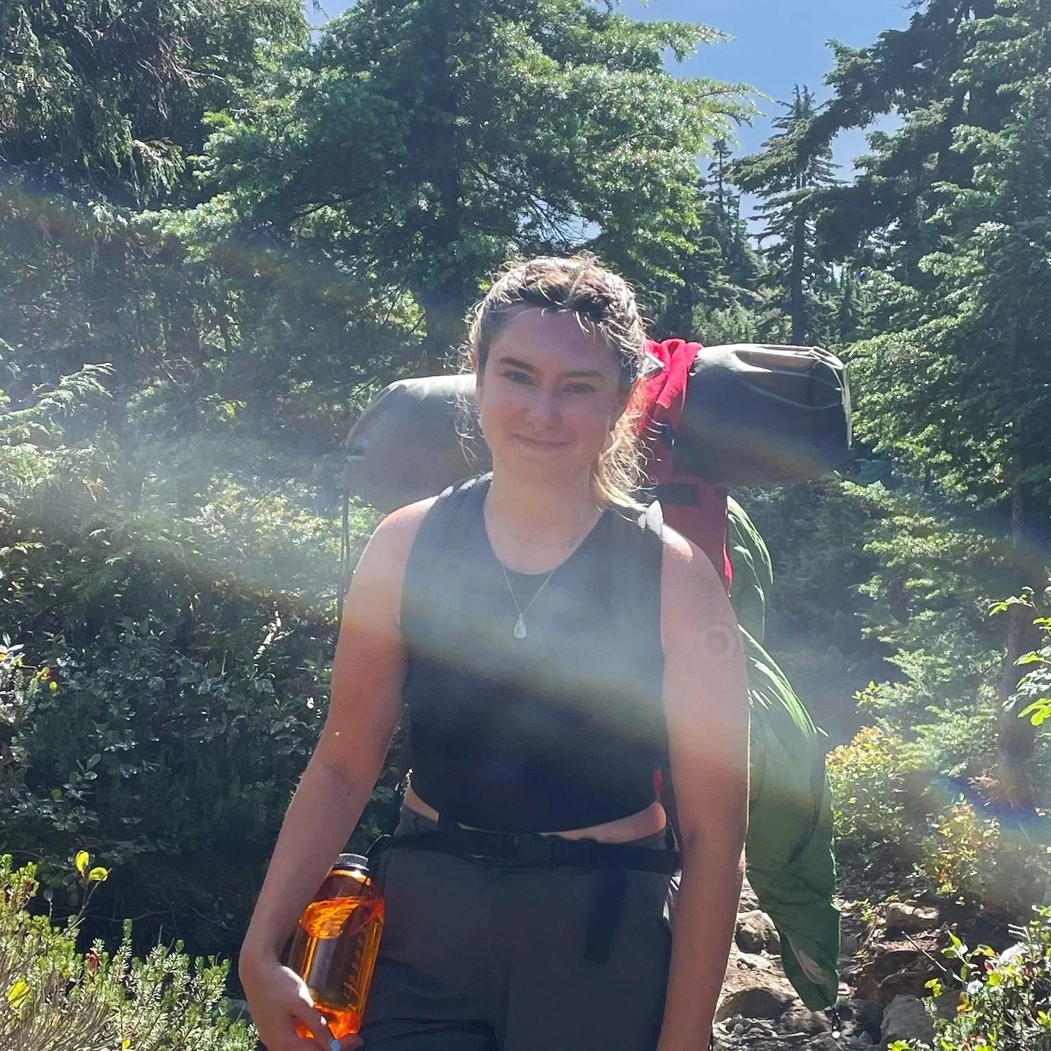 A woman hiking in a forest, wearing a black tank top, carrying a large backpack, and holding a water bottle, with trees and sunlight in the background.