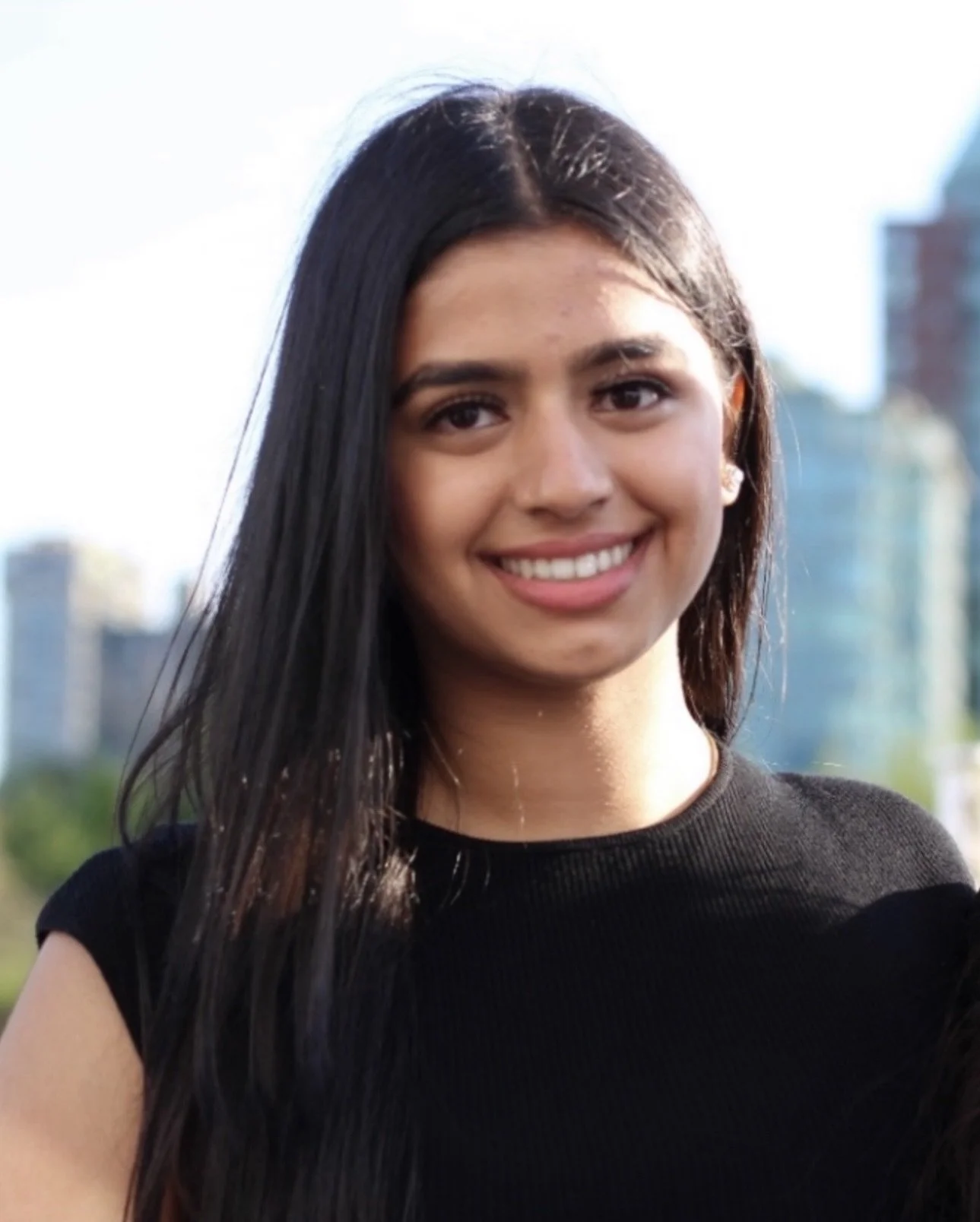 Close-up of a smiling young woman with long dark hair smiling outdoors with city buildings and trees in the background.