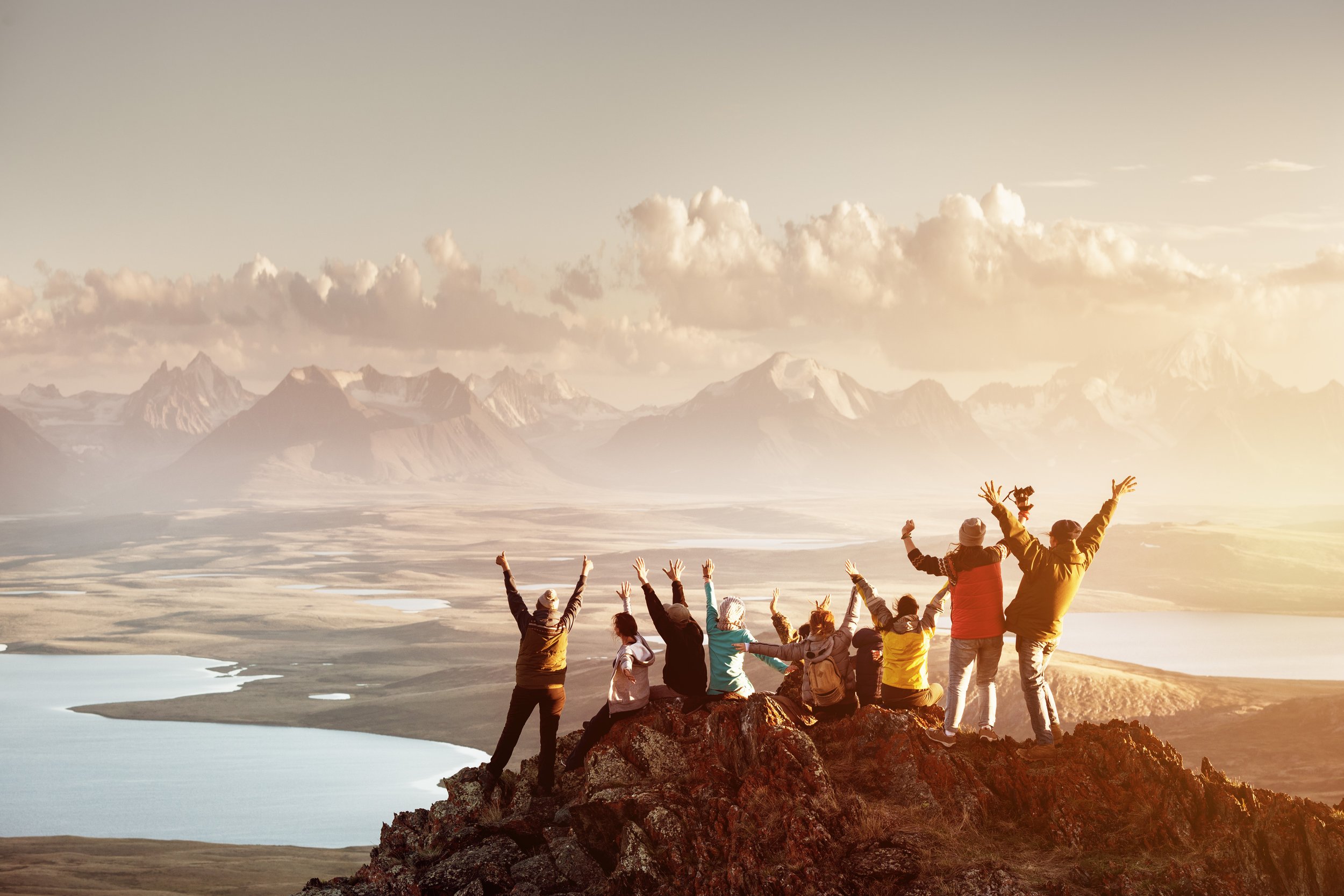 Group of people raising their hands on a rocky outcrop overlooking mountains and a valley with a river during sunset.