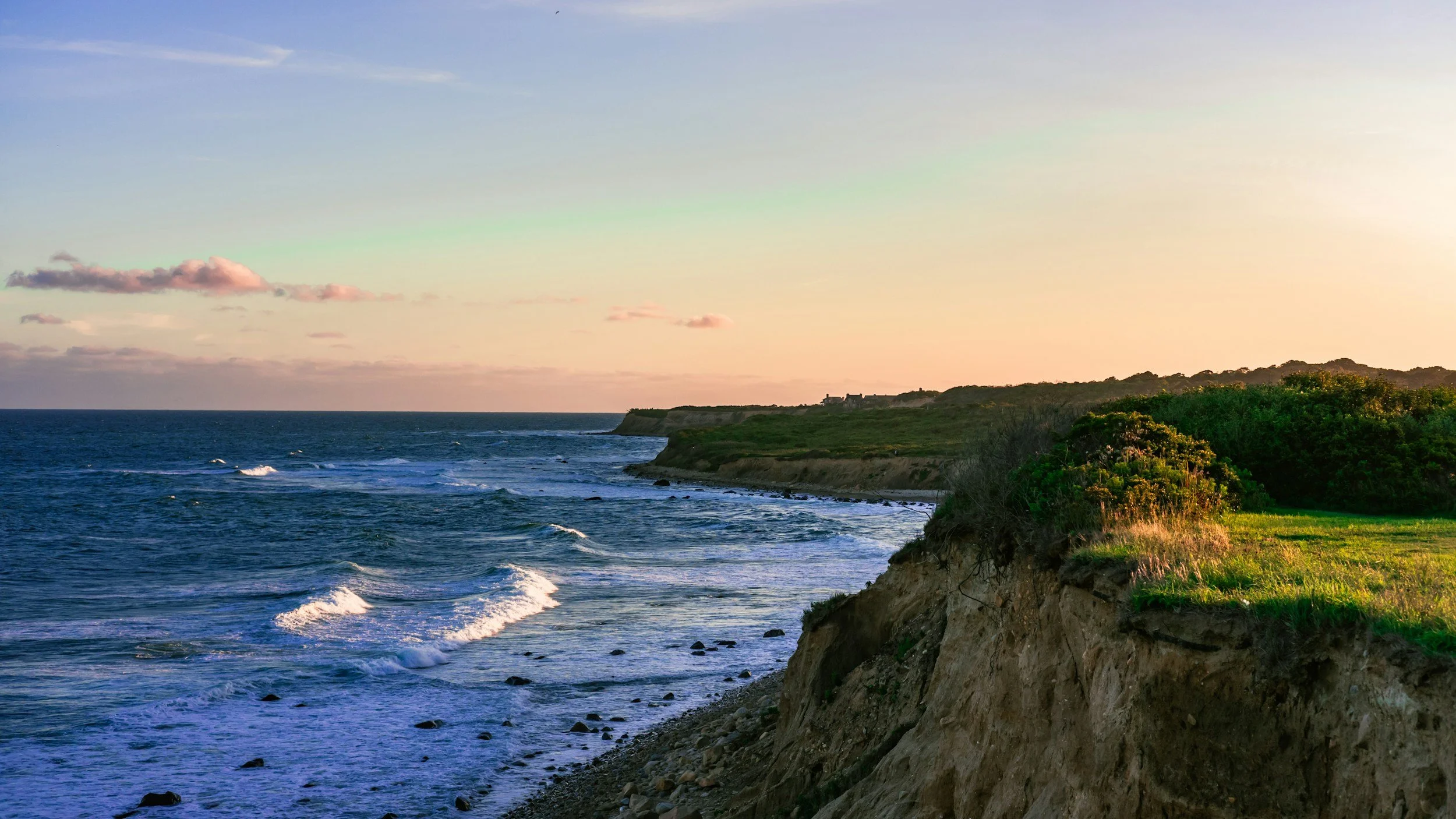 A scenic coastal landscape at sunset showing the ocean with waves crashing against rocky shoreline and a green hill with some houses in the distance.